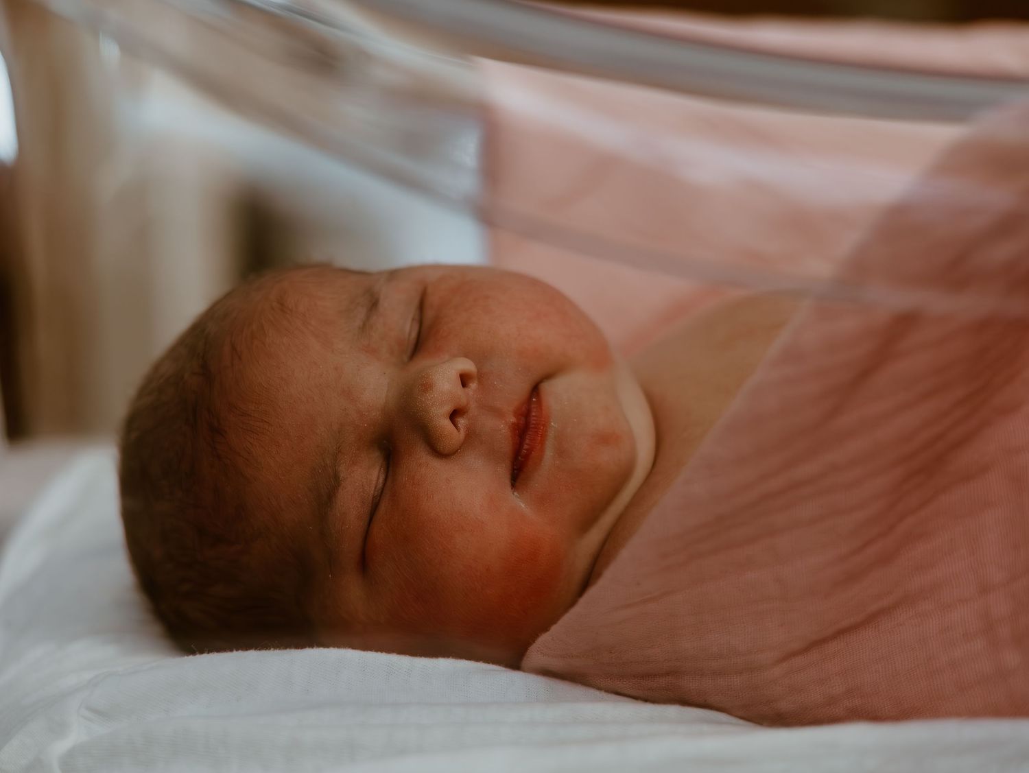 A peaceful sleeping newborn baby rests on soft pink bedding in warm lighting.