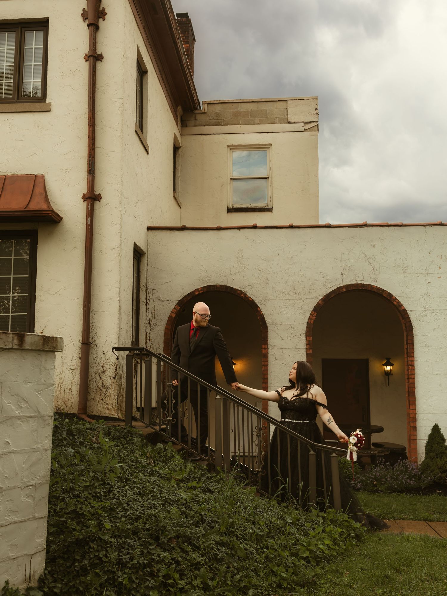 Couple in formal attire on stone steps of white stucco building with arched doorways under cloudy sky.