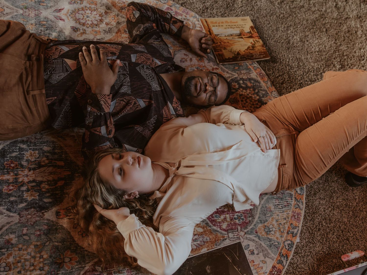 Two people lying on a patterned rug, one in a white top and tan pants, the other in a colorful jacket.