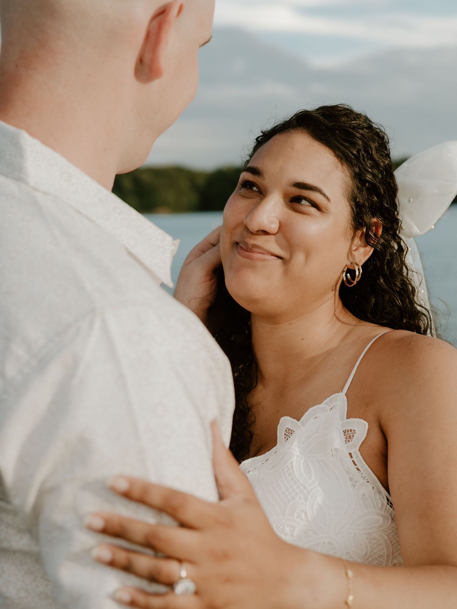 Intimate wedding couple embraces against cloudy sky and waterfront backdrop during romantic outdoor photoshoot.