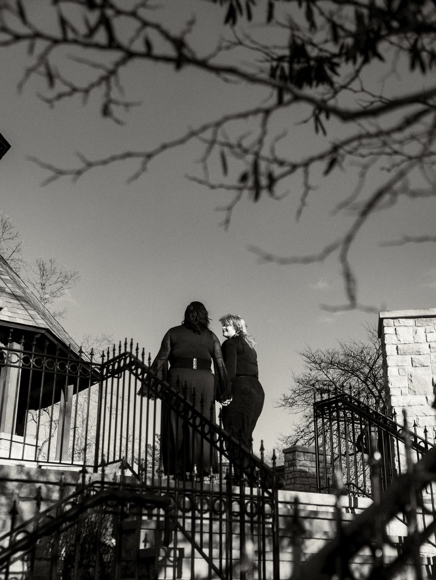 Two silhouettes stand against an iron gate at dusk, creating a dramatic black and white composition.