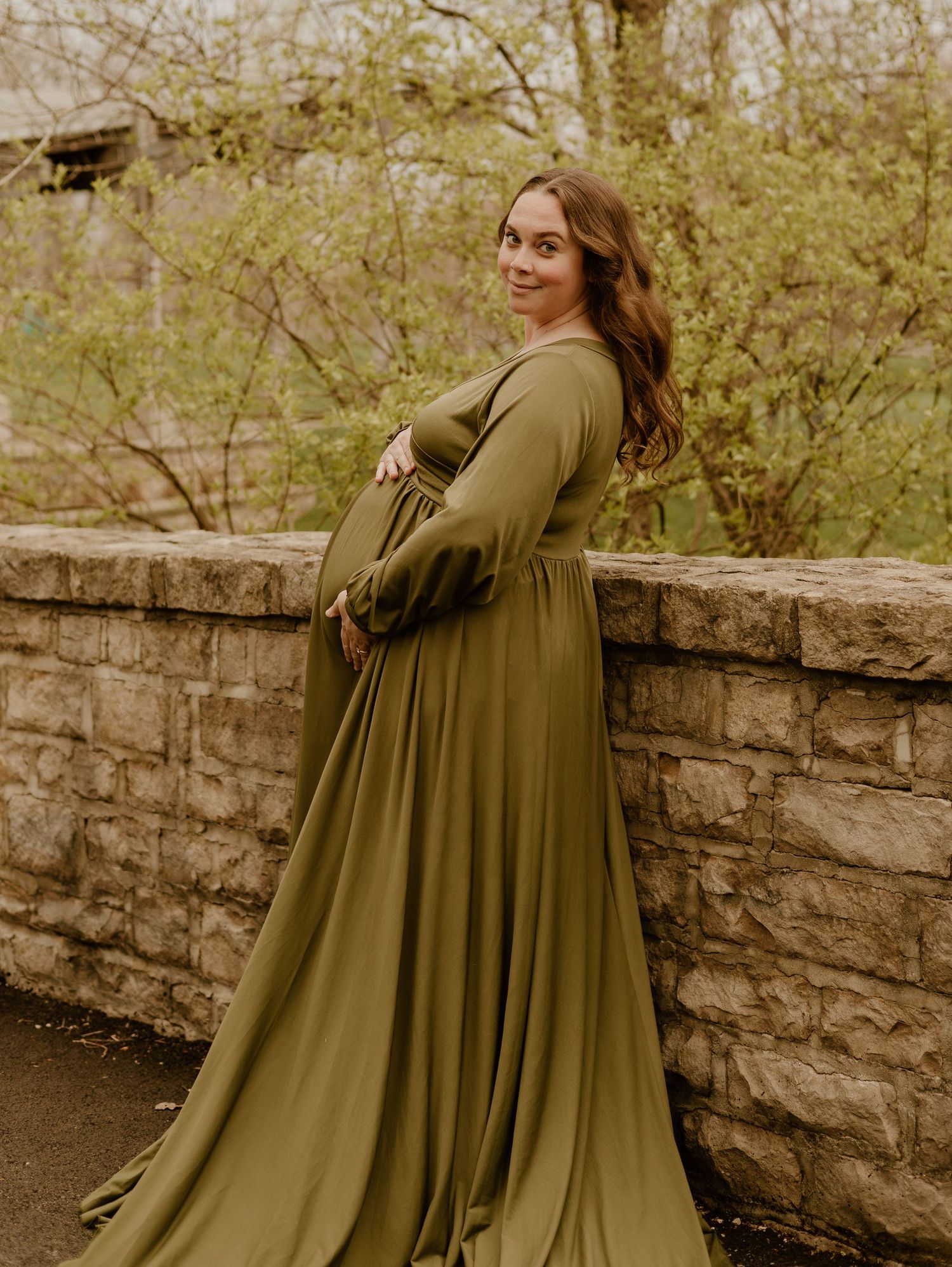 Woman in olive green flowy dress poses against stone wall in autumn outdoor setting.