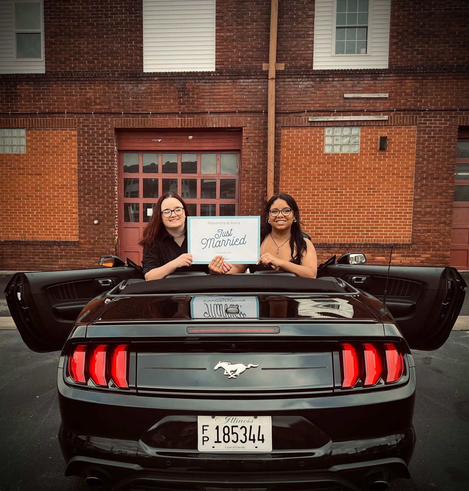 Modern Mustang convertible parked in front of brick building with a sign being held up.