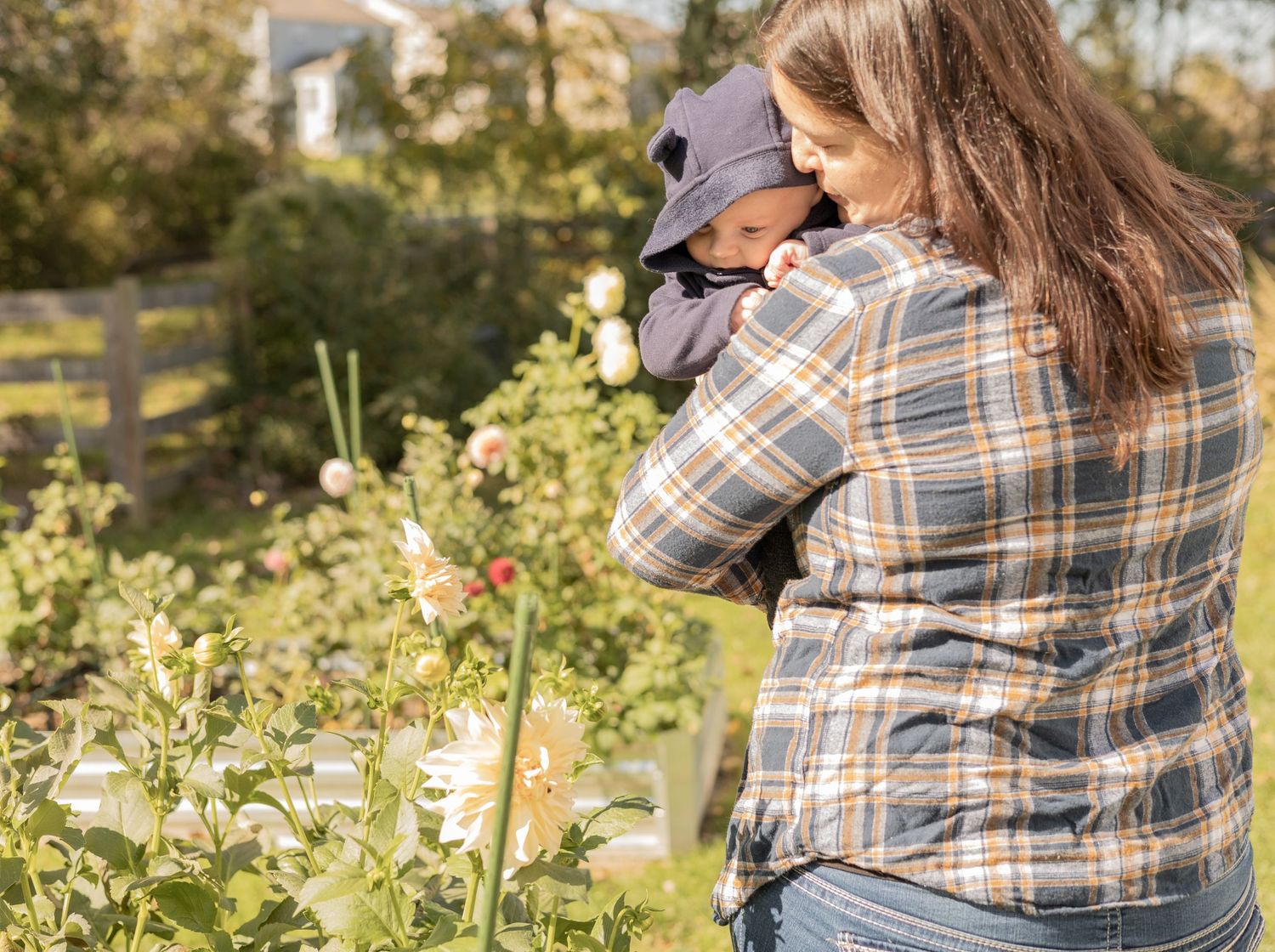 Person wearing plaid flannel hugging a baby while standing in a garden with blooming flowers and greenery.