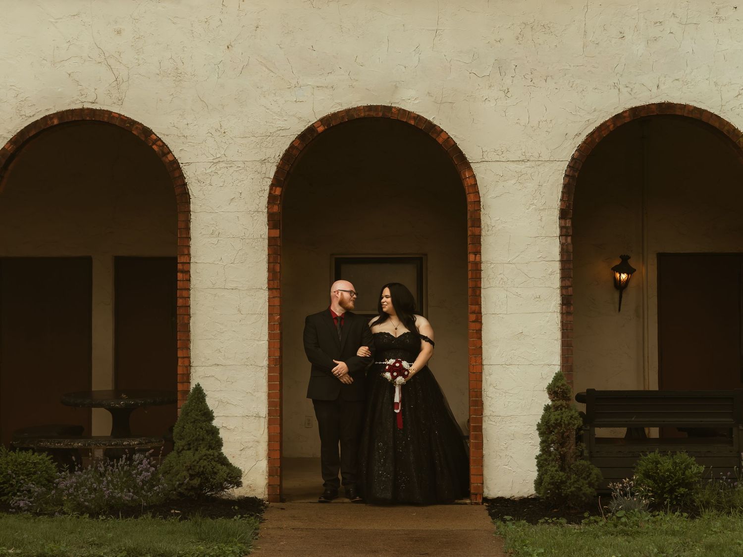 Couple in matching black wedding attire posing under brick arched portico of white stucco building.