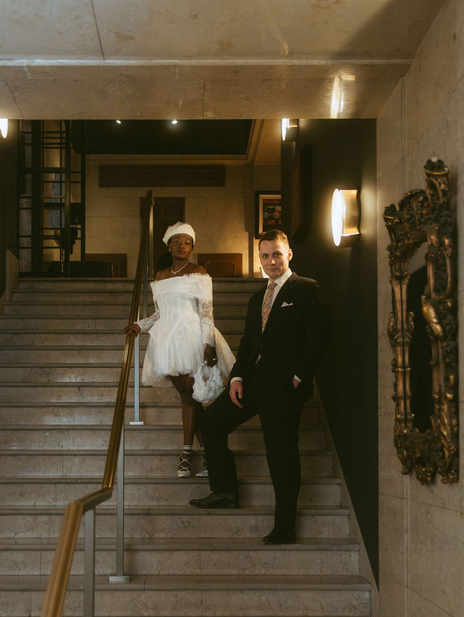 Couple in wedding attire on marble stairs, bride in short white dress and groom in dark suit, elegant hotel interior.