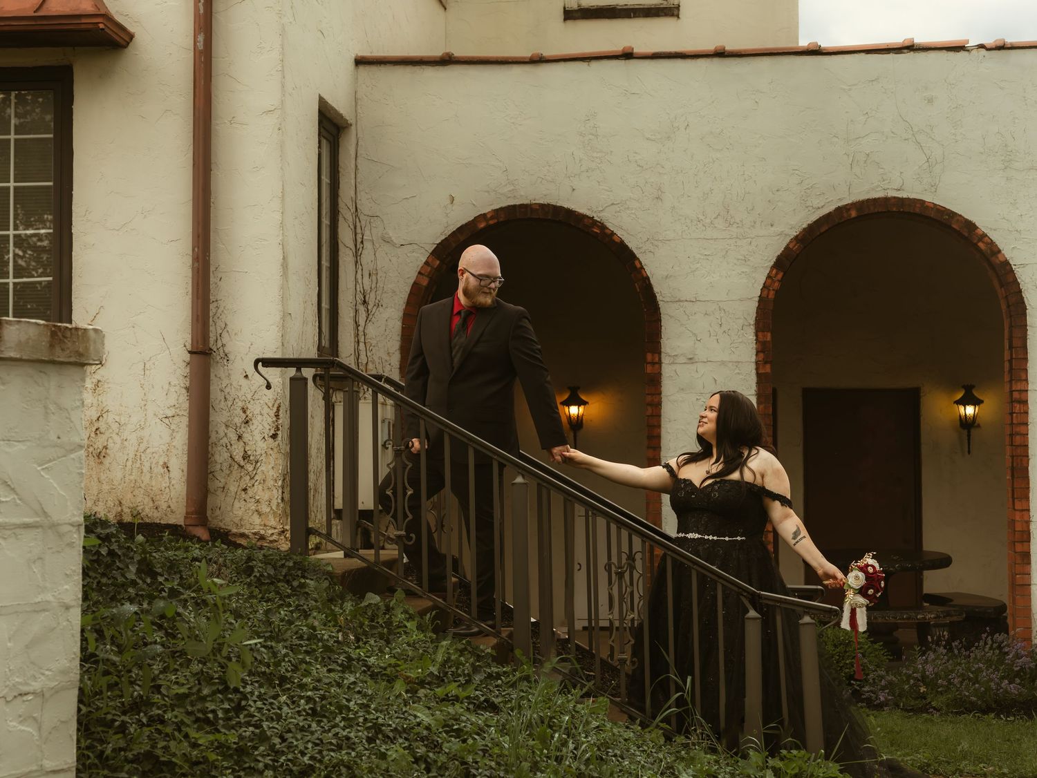Couple holding hands on outdoor staircase of historic building with arched brick doorways and lush greenery.