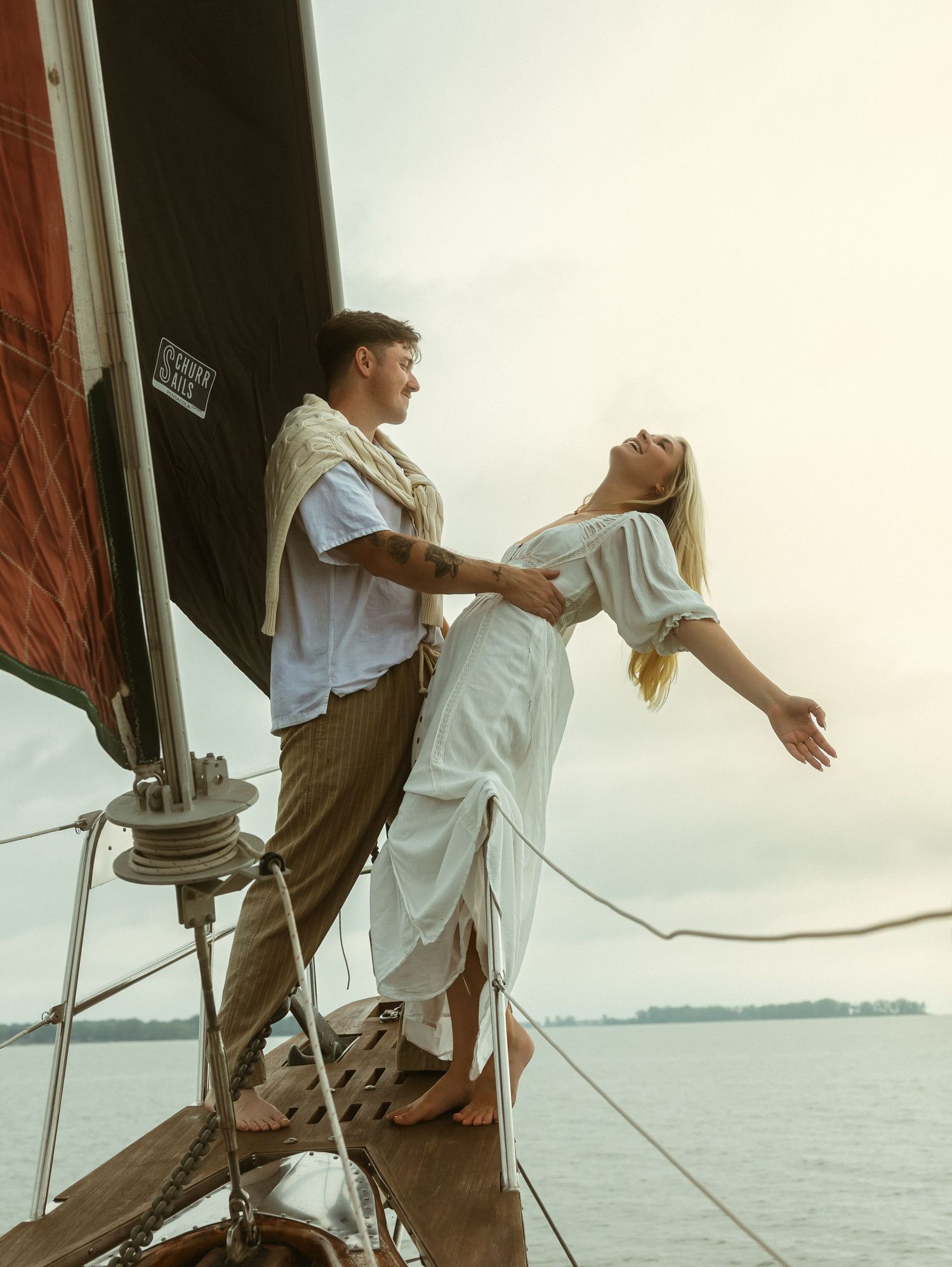A couple stands on a sailboat bow, woman in white dress leaning back with arms open, man holding her over open water.