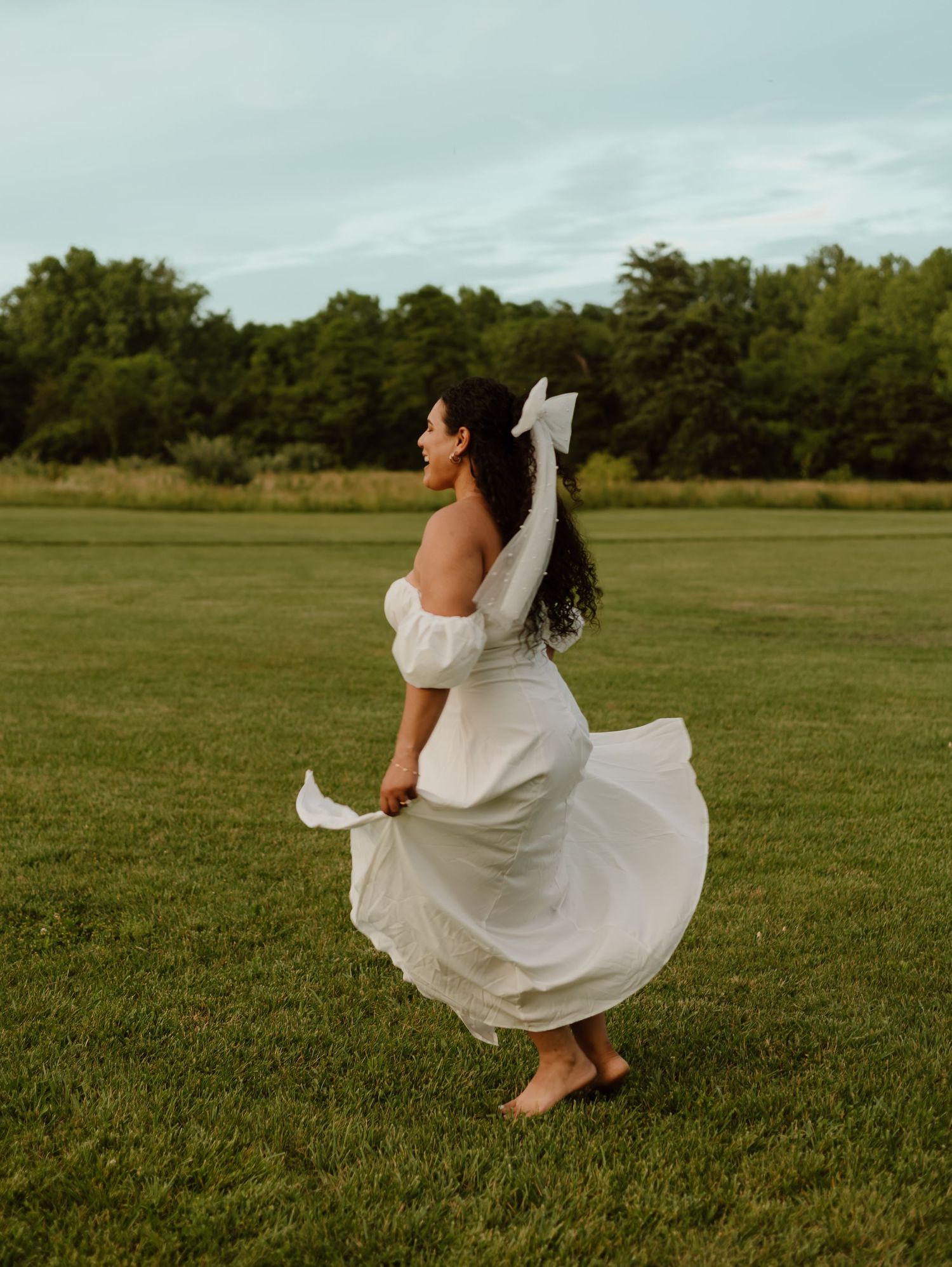 A person in a flowing white dress twirls gracefully across a green grassy field on a cloudy day.
