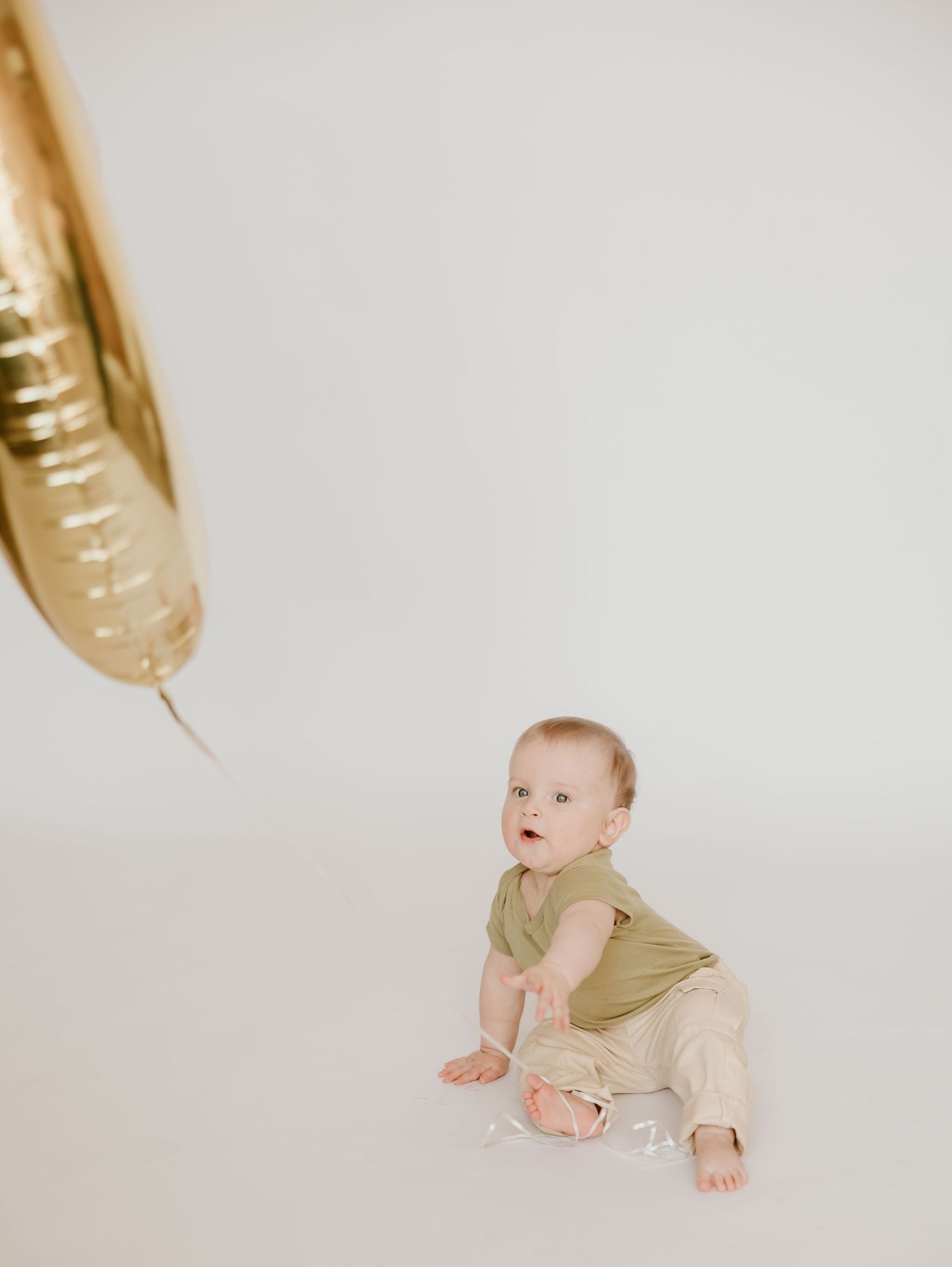 A baby in beige clothing crawls on a white background while gold balloons float above.