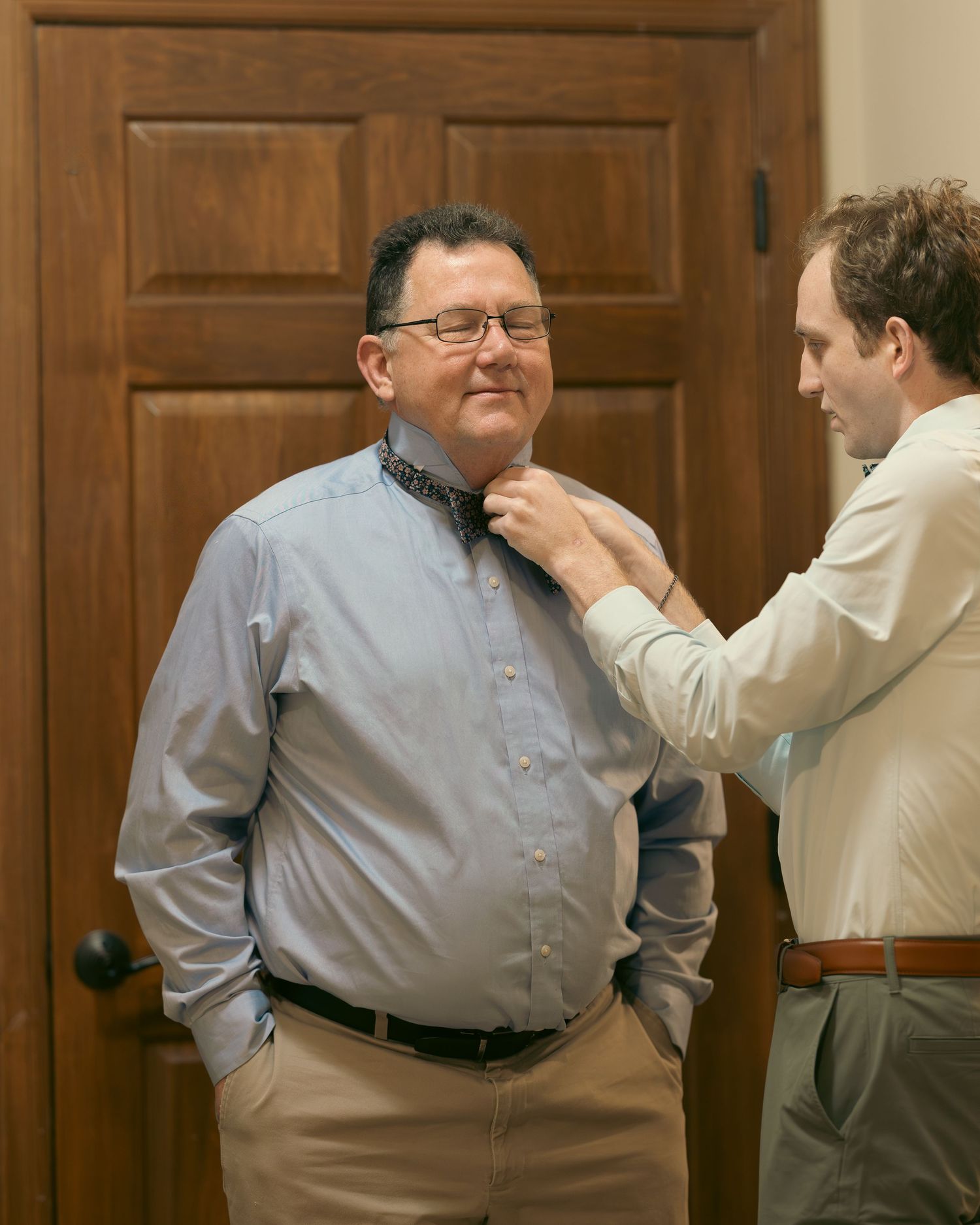 A person helps adjust another person's tie while standing in front of a wooden door.