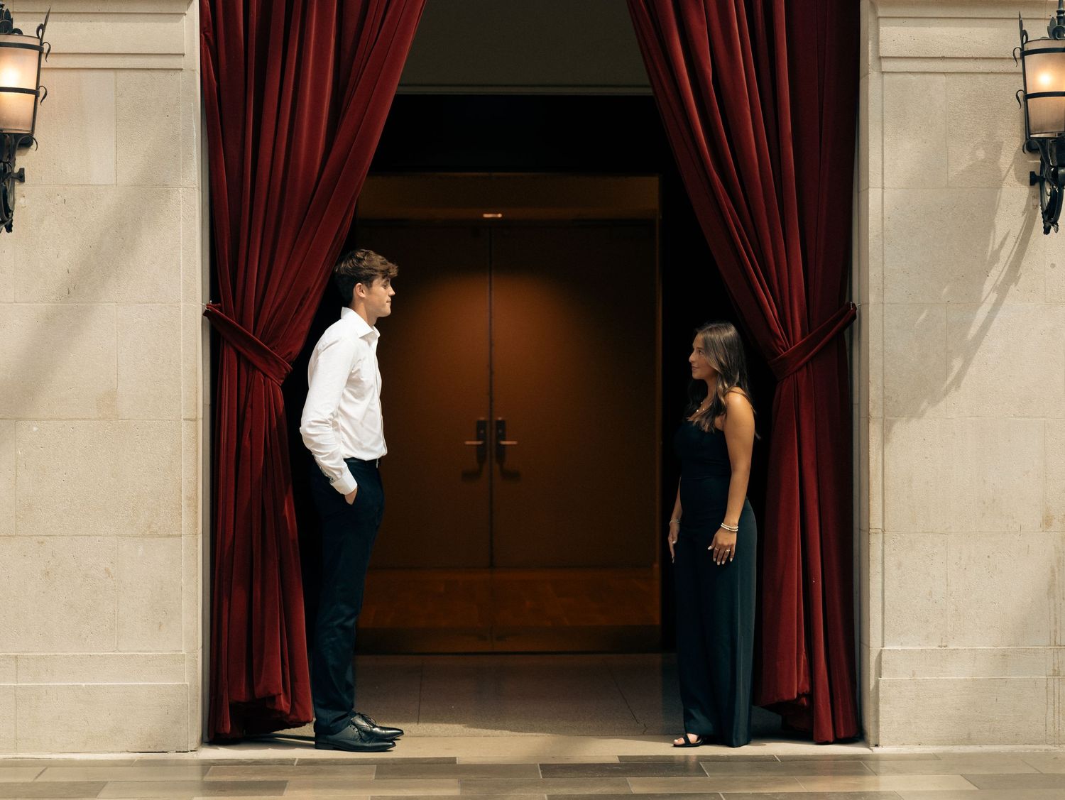 Silhouettes stand near burgundy curtains in a dramatic hallway setting.