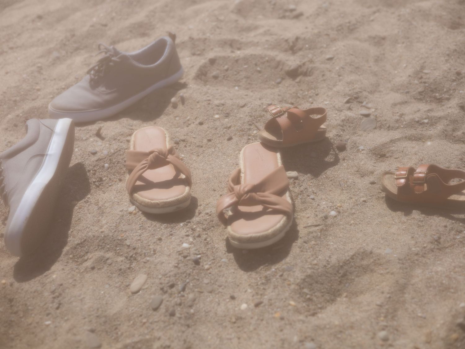 Multiple pairs of sandals and bare feet stand in a line on sandy ground at the beach.