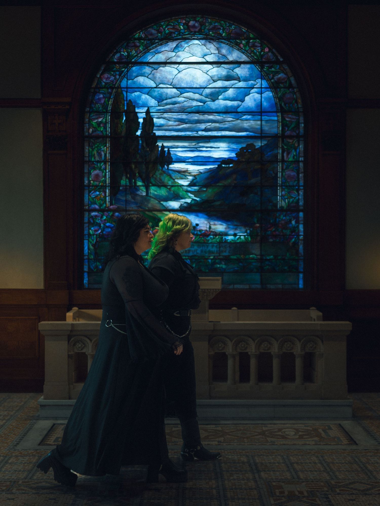 Silhouetted figures stand against a glowing stained glass window depicting a moonlit landscape at night.