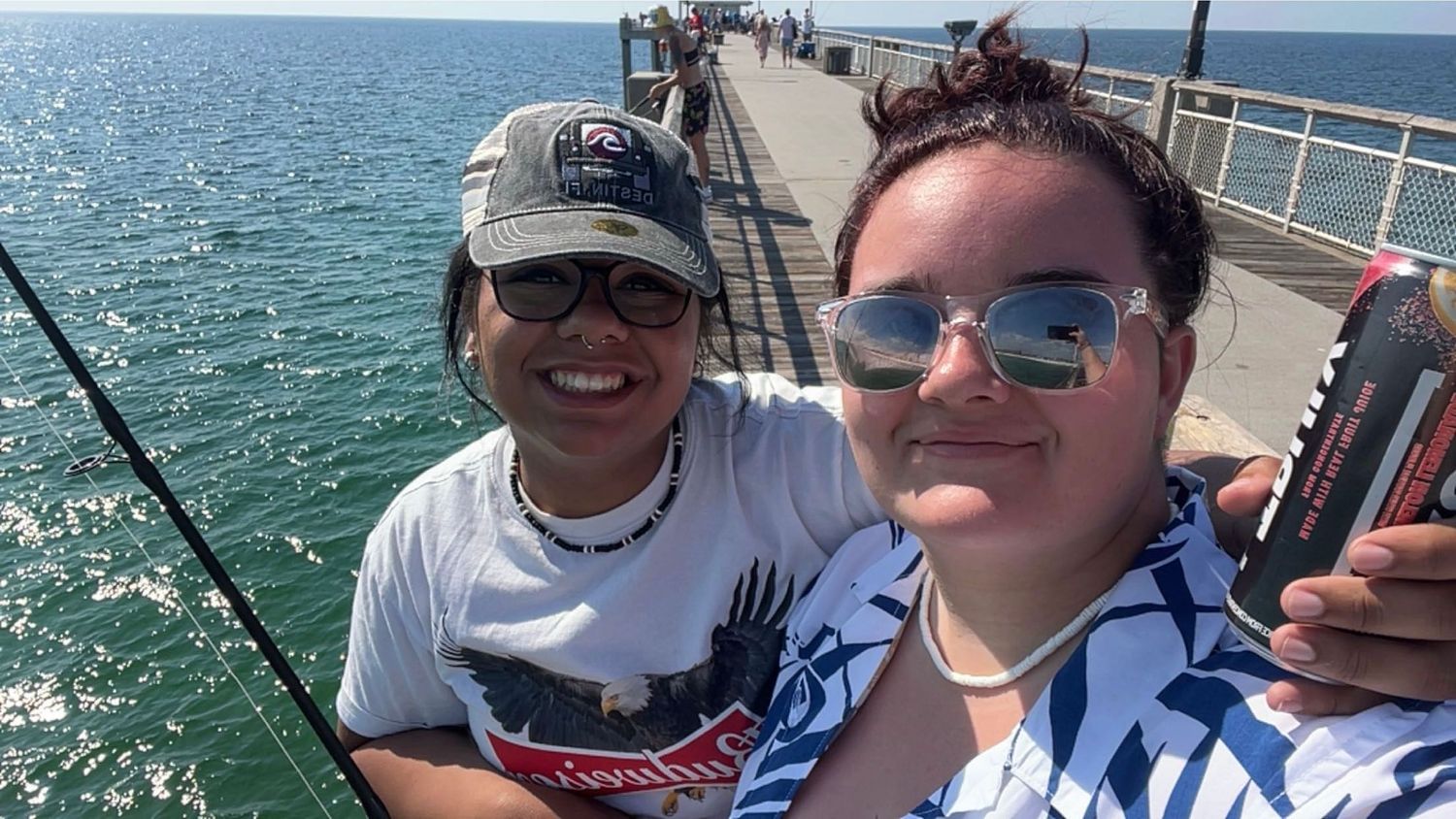 Two people taking a selfie on a sunny pier with ocean views in the background.