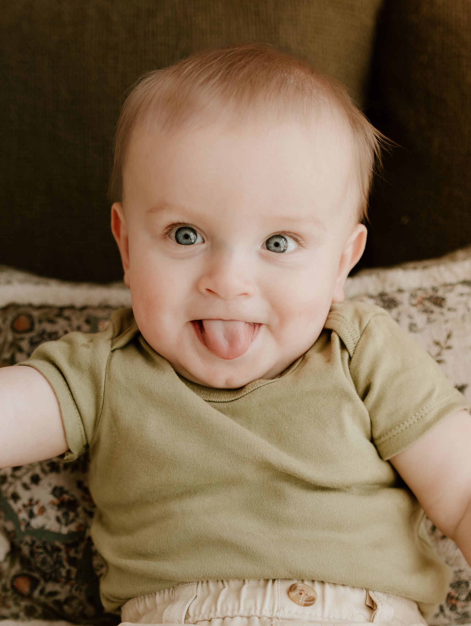 A baby in a beige onesie lies on patterned fabric showing four different playful expressions and sticking out tongue.