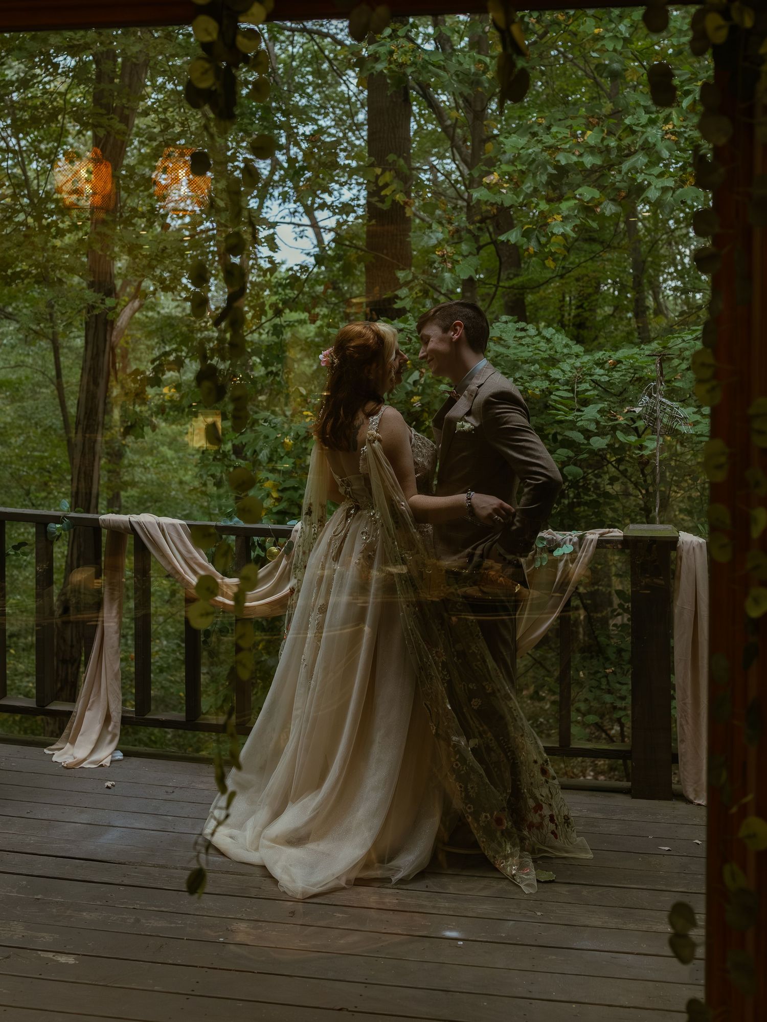 A couple shares an intimate moment on a forest deck decorated with hanging lights at dusk.