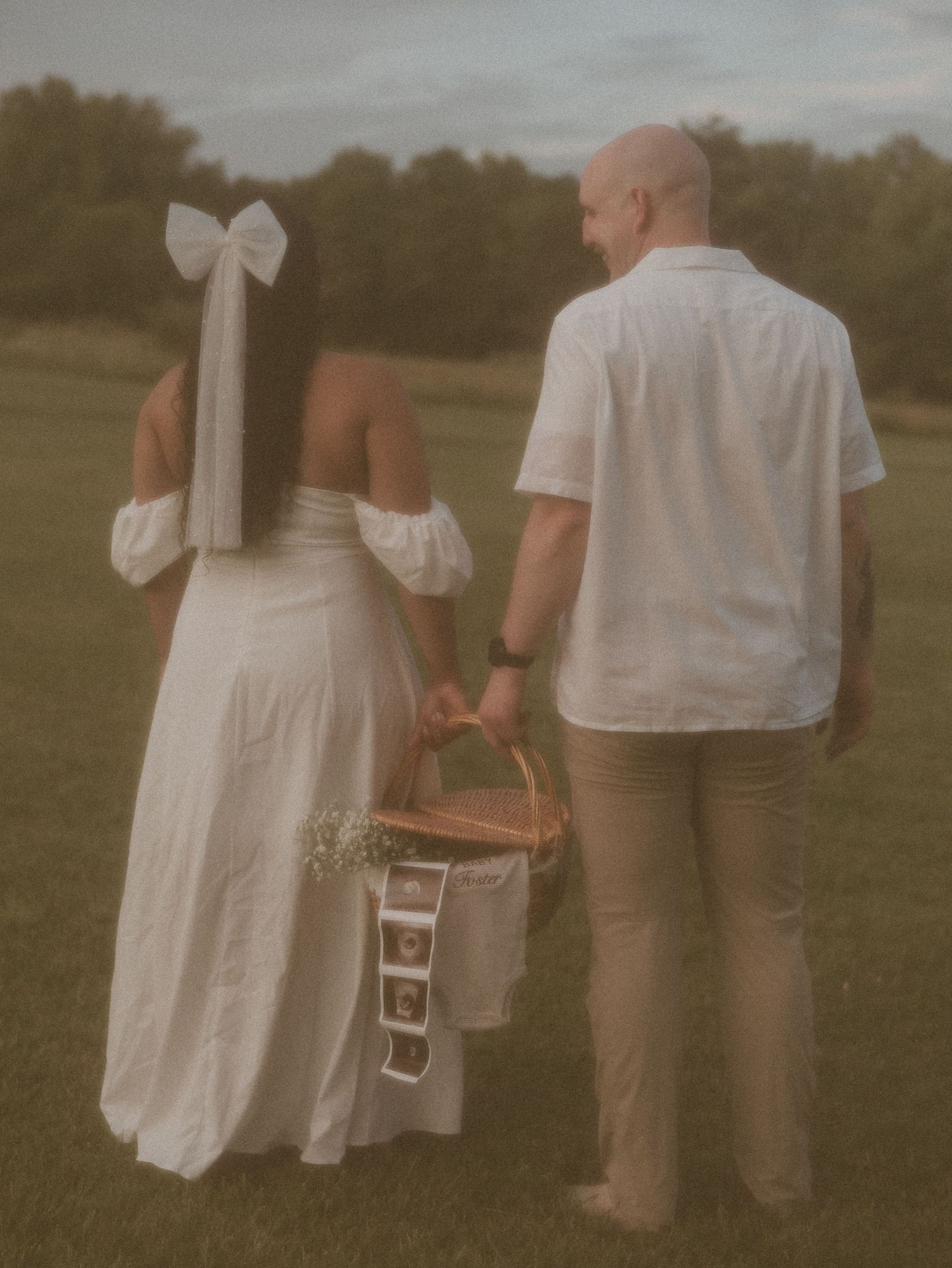 A couple in wedding attire holding hands and carrying a picnic basket together in a grassy field at dusk.
