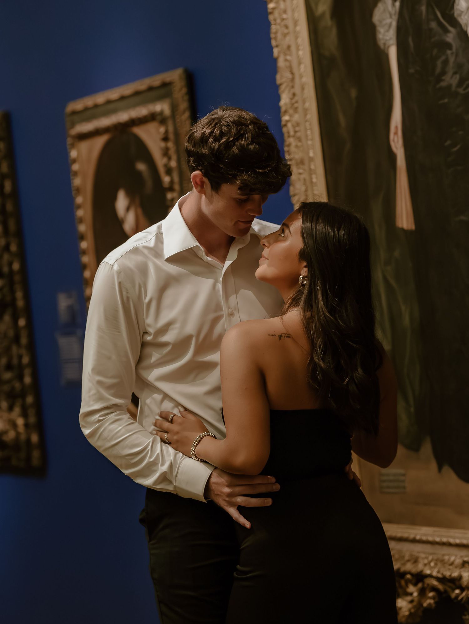 A romantic couple embraces and shares a tender kiss in an elegant indoor setting with ornate frames on the wall.