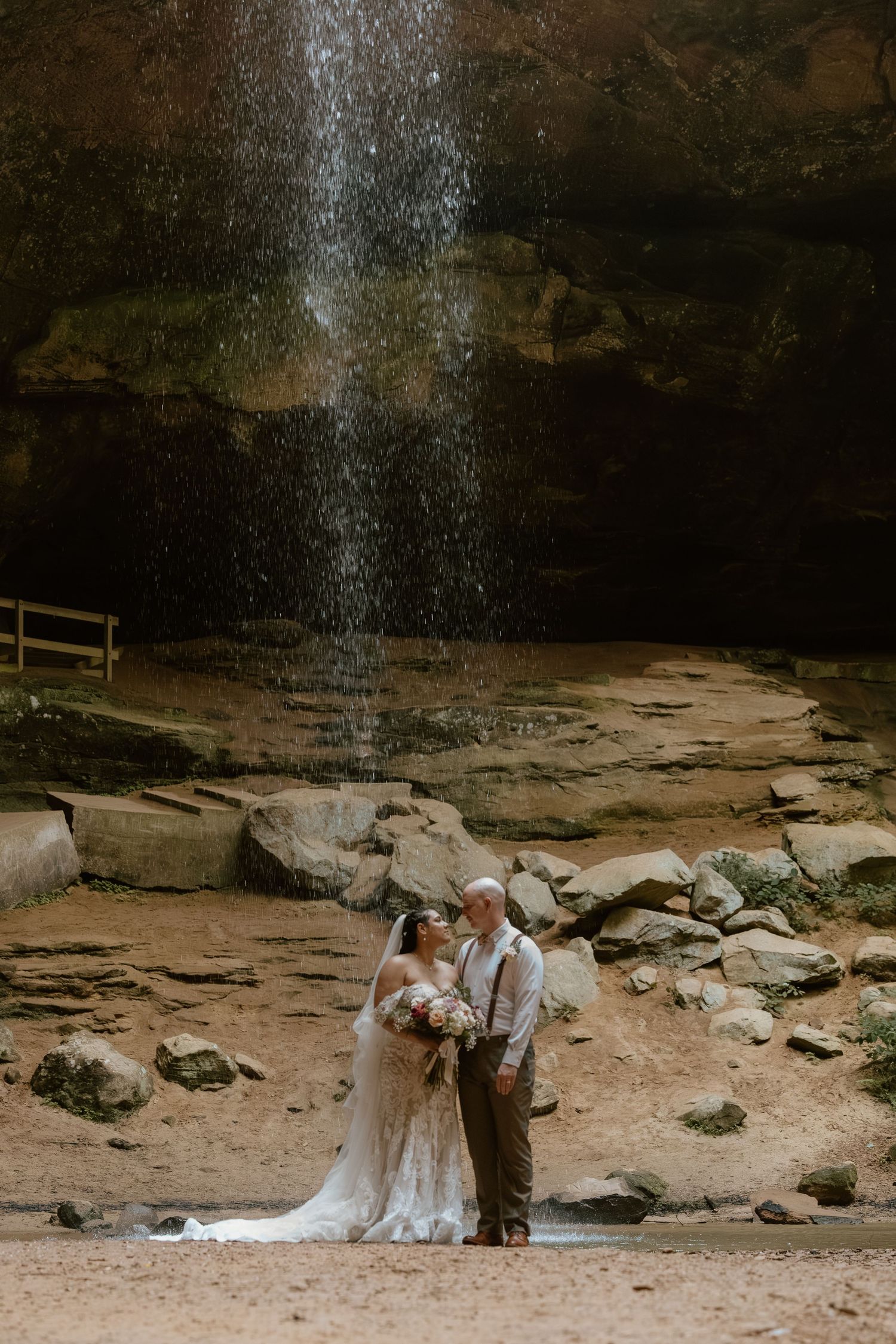 A couple shares an intimate moment under a cascading waterfall in a rocky cave setting.
