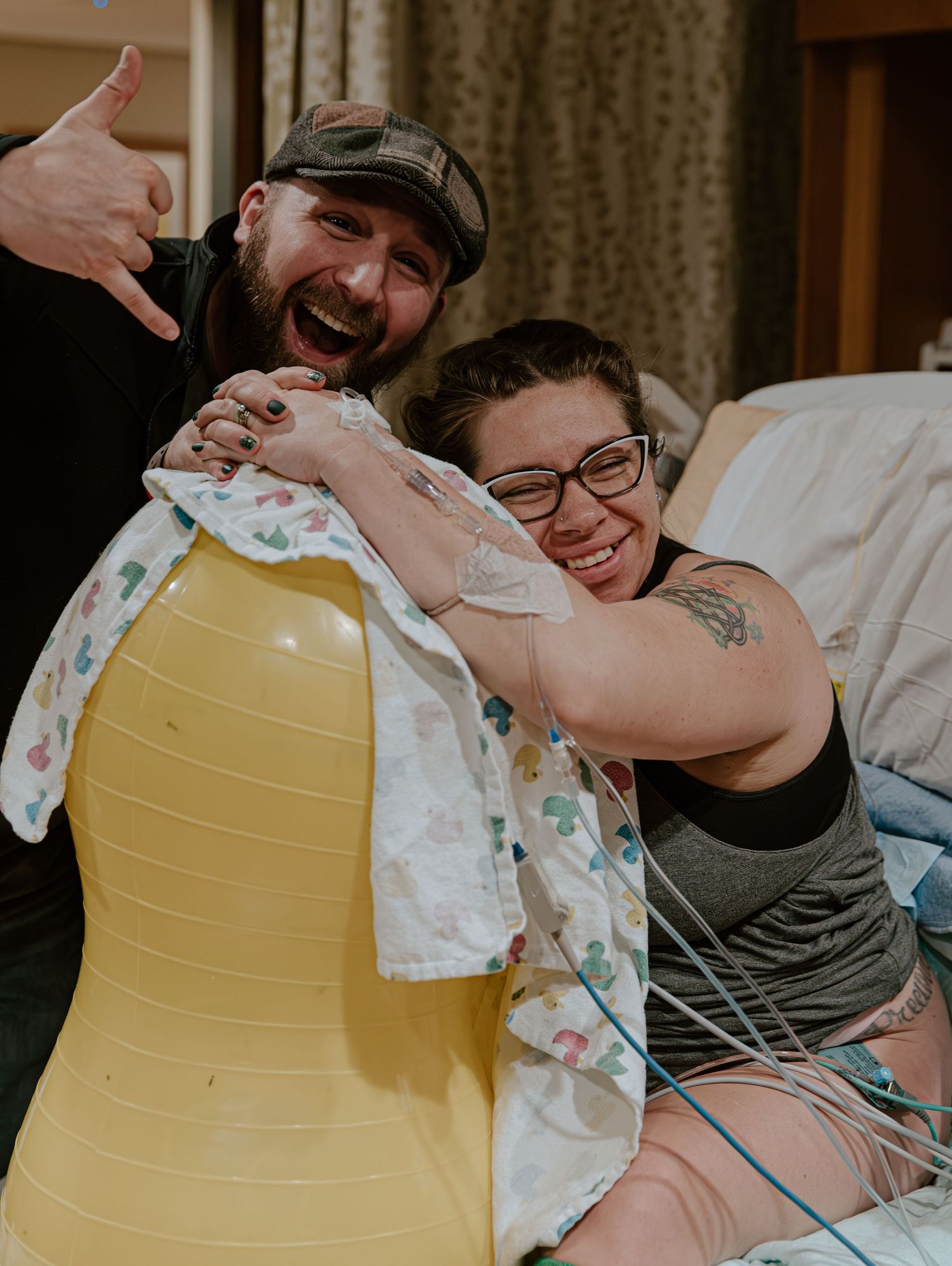 Couple smiling joyfully in hospital room, woman leaning on yellow birthing ball during labor, man giving shaka sign.