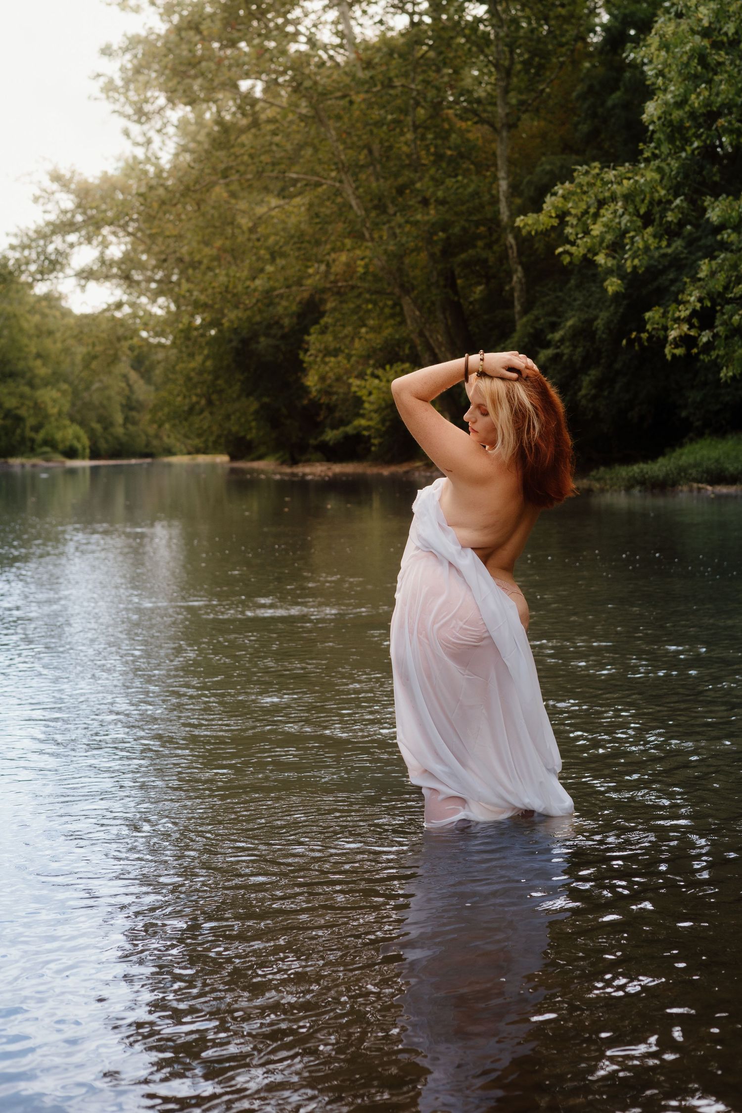 A figure in a white top wades through a tranquil river surrounded by green trees on a summer day.