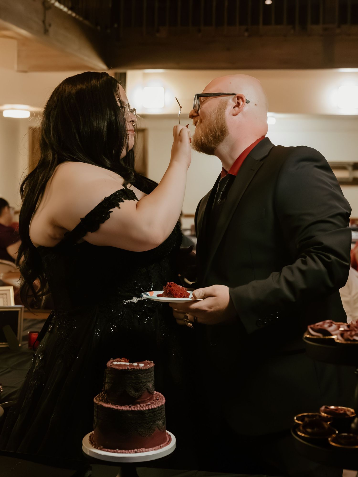 Bride in black gown feeds groom cake at gothic wedding with dark red tiered cake on table.