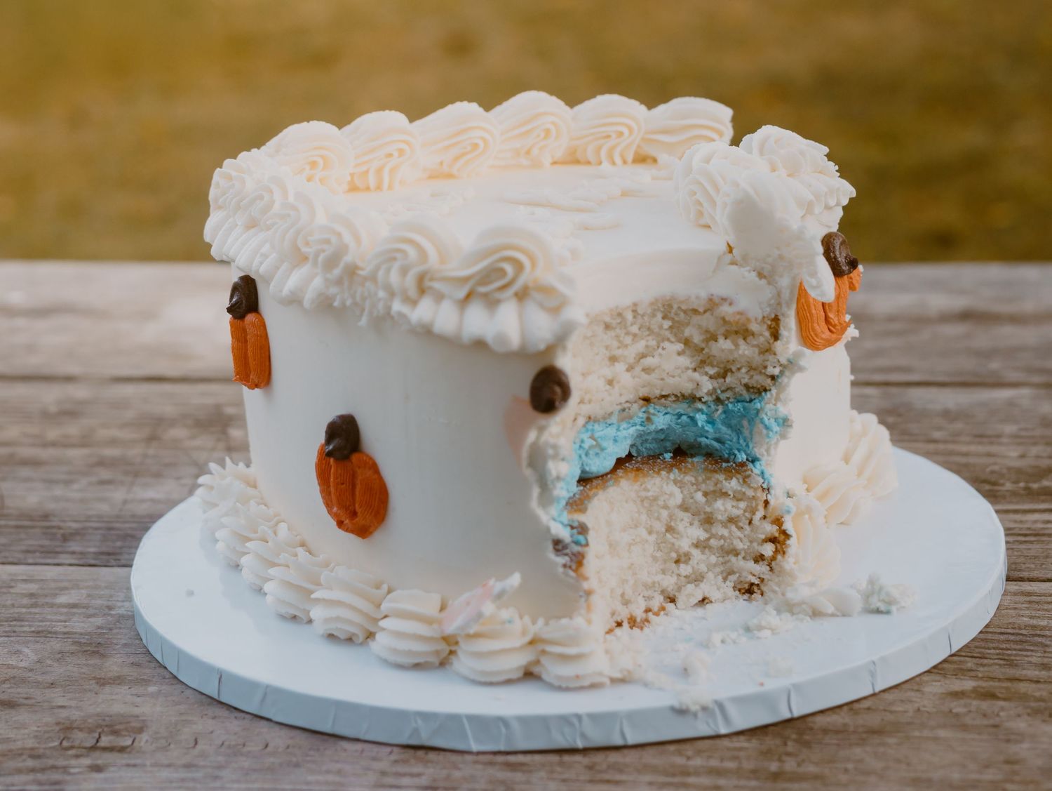 A white frosted cake with blue accents and decorative macarons sits on a rustic wooden surface.