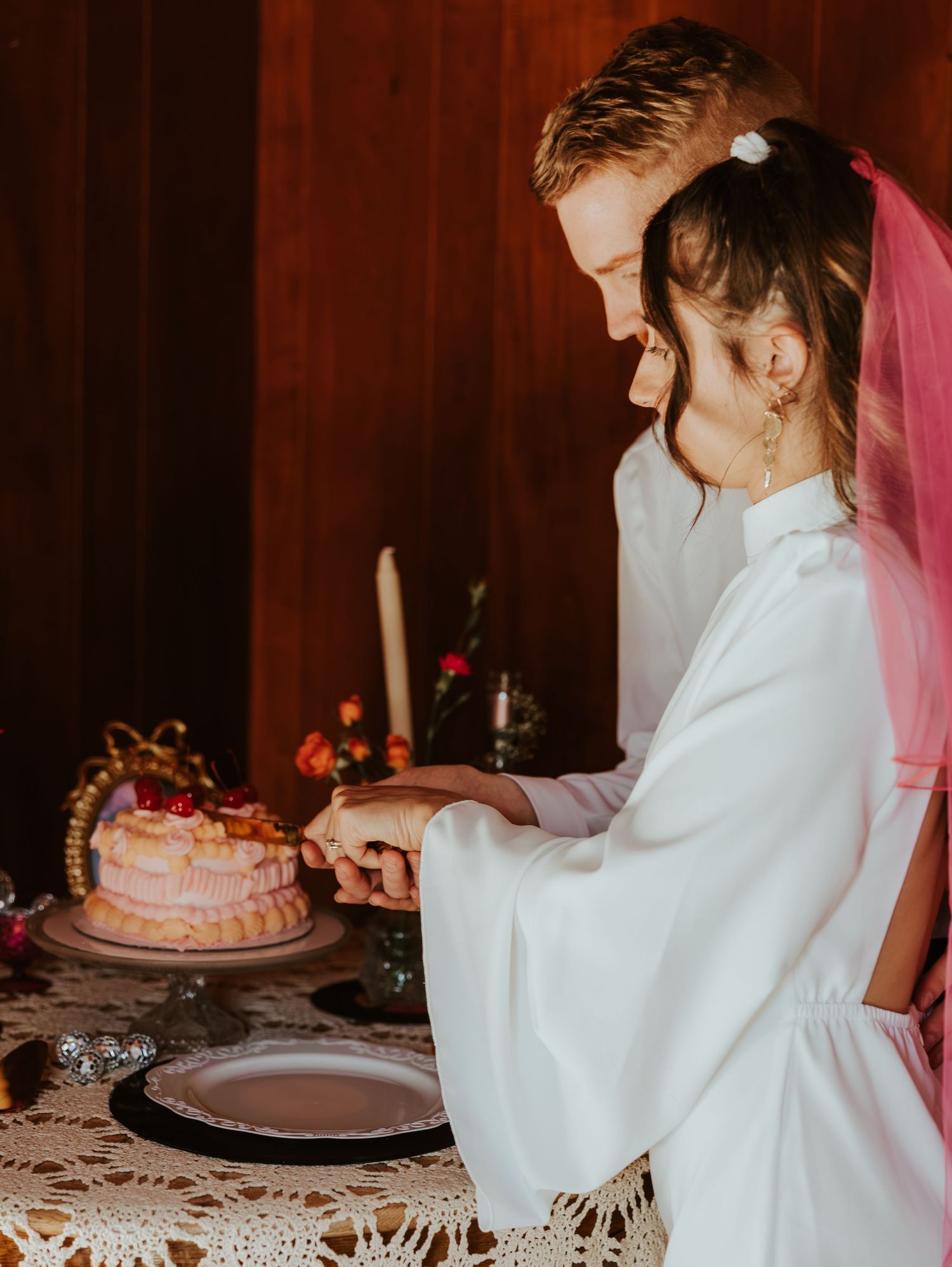 A bride and groom cut into a pink frosted wedding cake together at their candlelit reception.