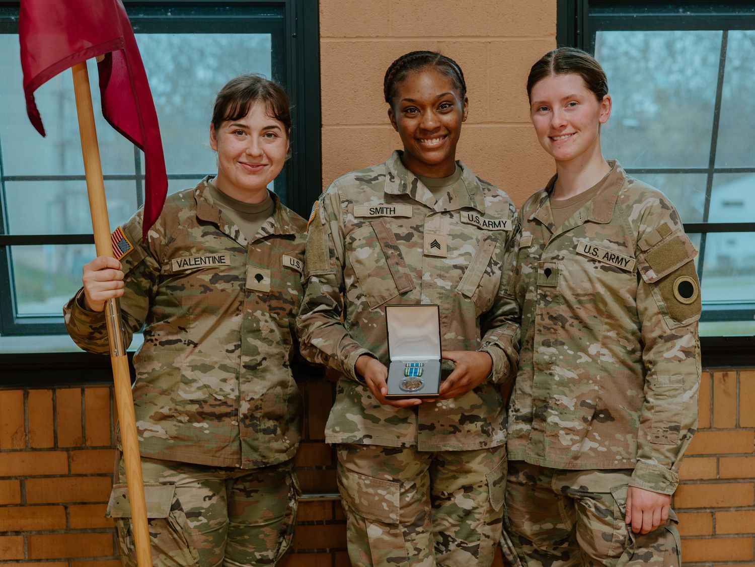 Three U.S. Army service members in camouflage uniforms pose with a plaque and red flag in an indoor setting.