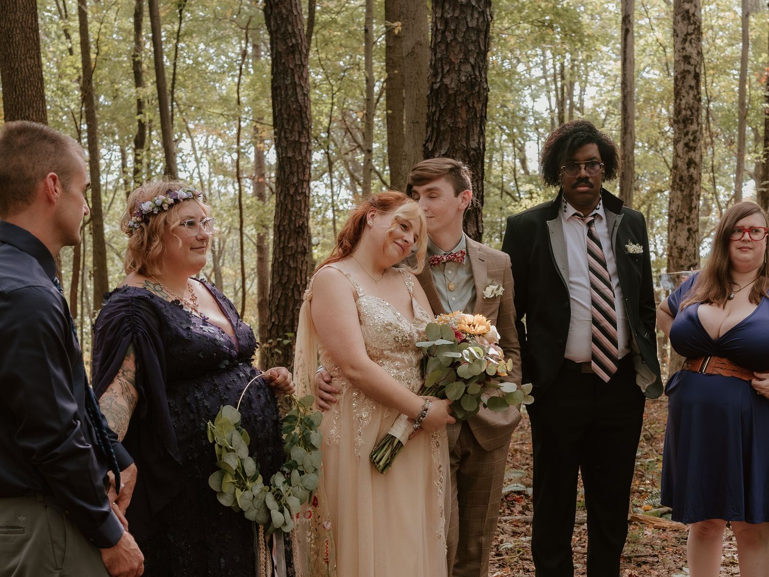 Wedding party members pose together in a sunlit forest setting wearing formal attire and holding bouquets.