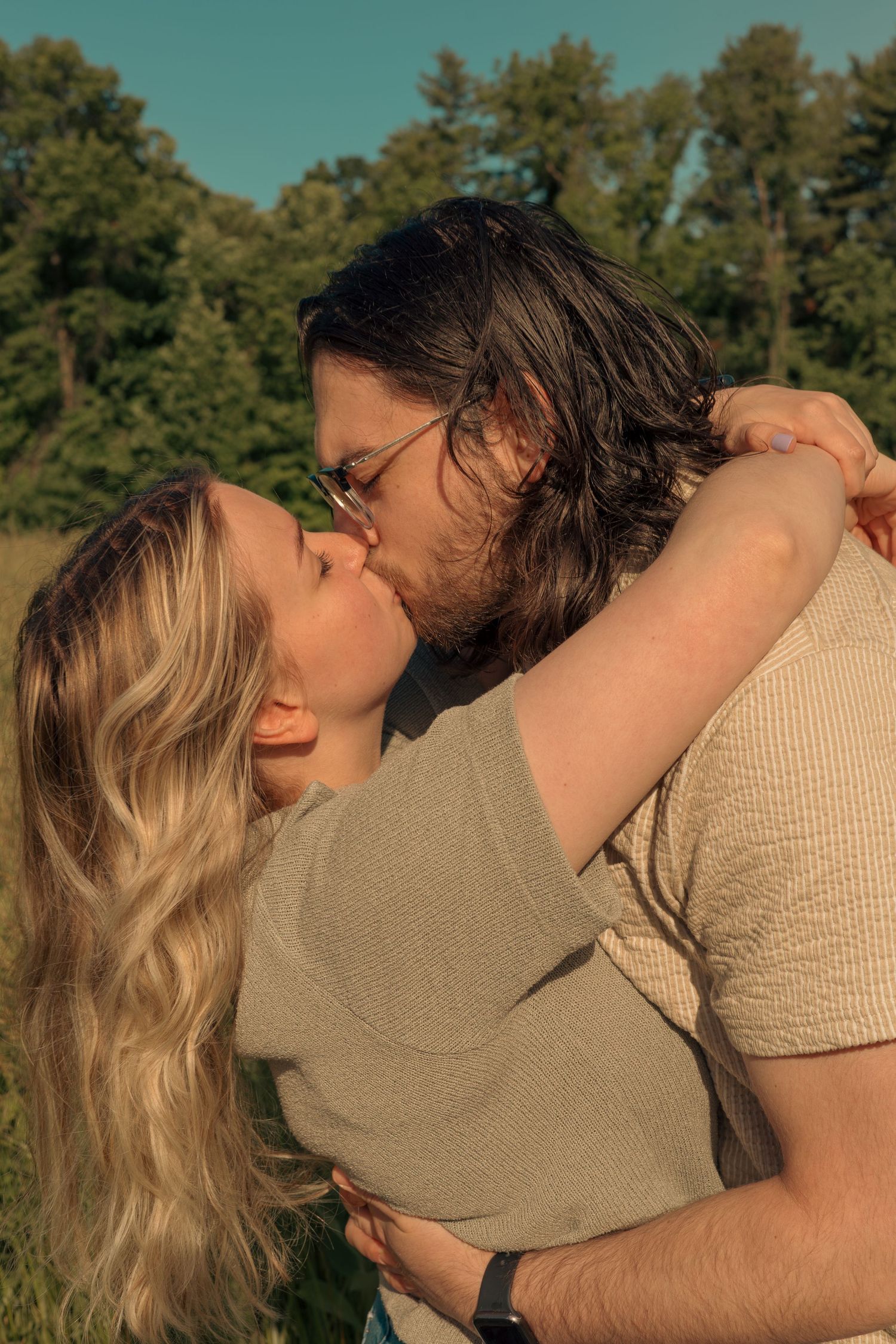 A romantic couple sharing an intimate kiss outdoors with sunlit pine trees in the background.