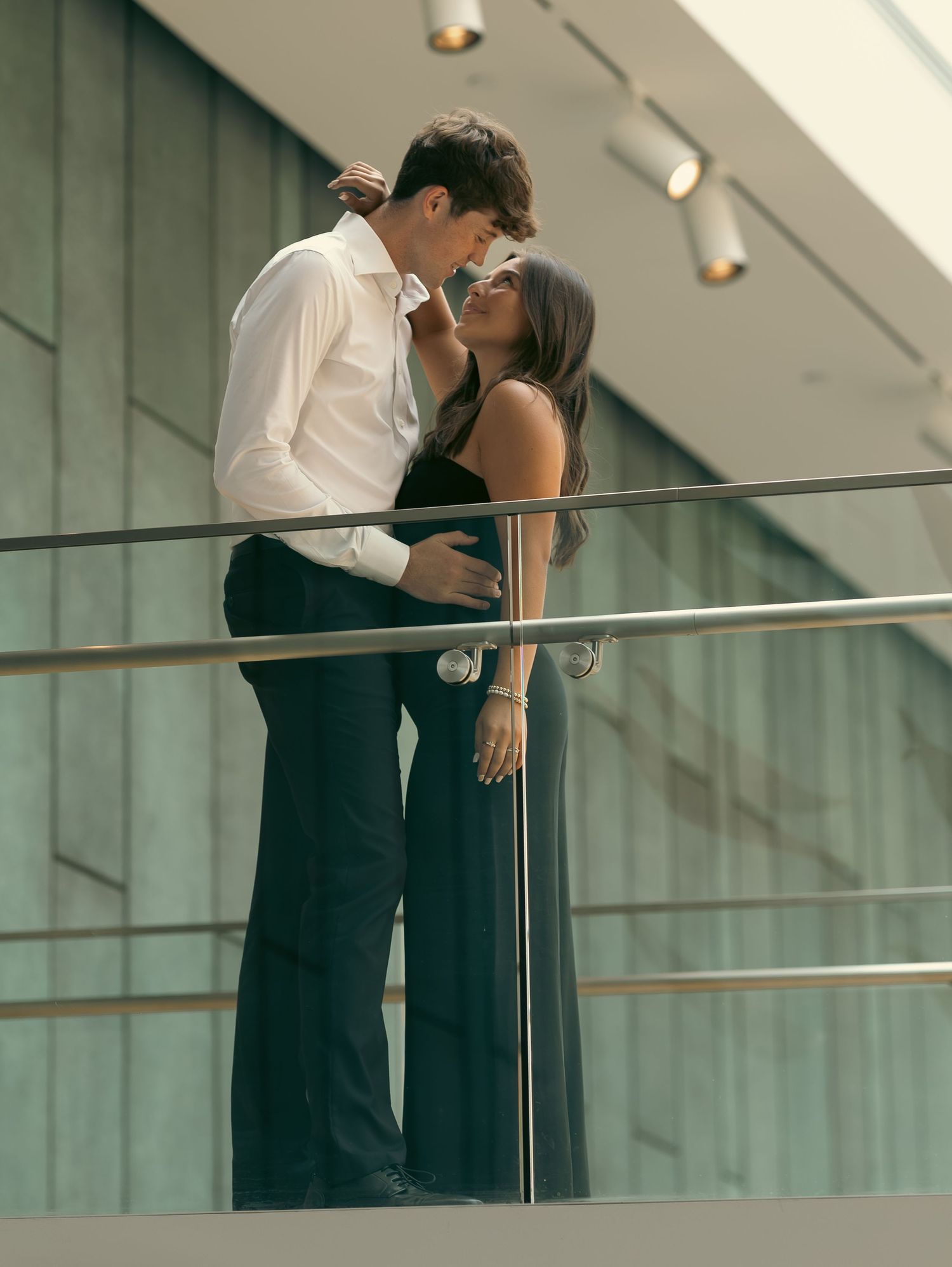 A romantic couple in formal wear share an intimate moment by a modern glass railing in an indoor setting.