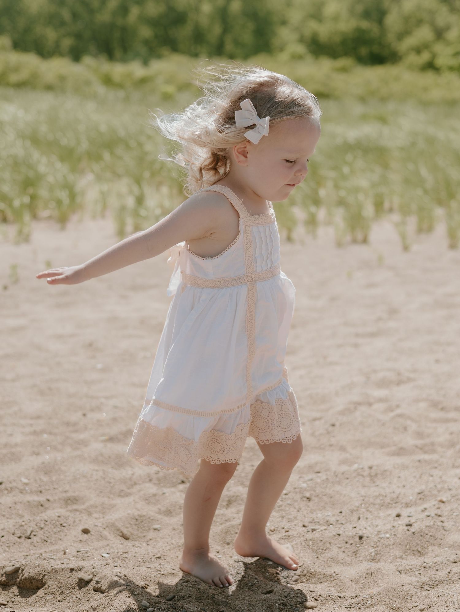 Young child in white summer dress twirls and plays on sandy beach in soft sunlit meadow setting.