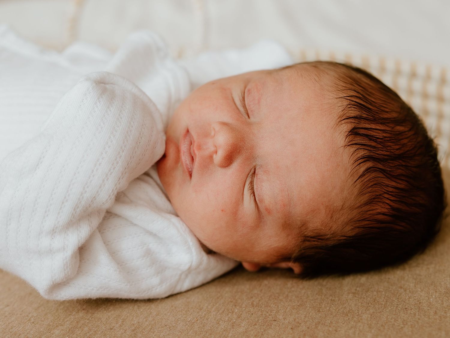 A sleeping newborn baby wrapped in a soft white swaddle blanket rests peacefully on beige fabric.