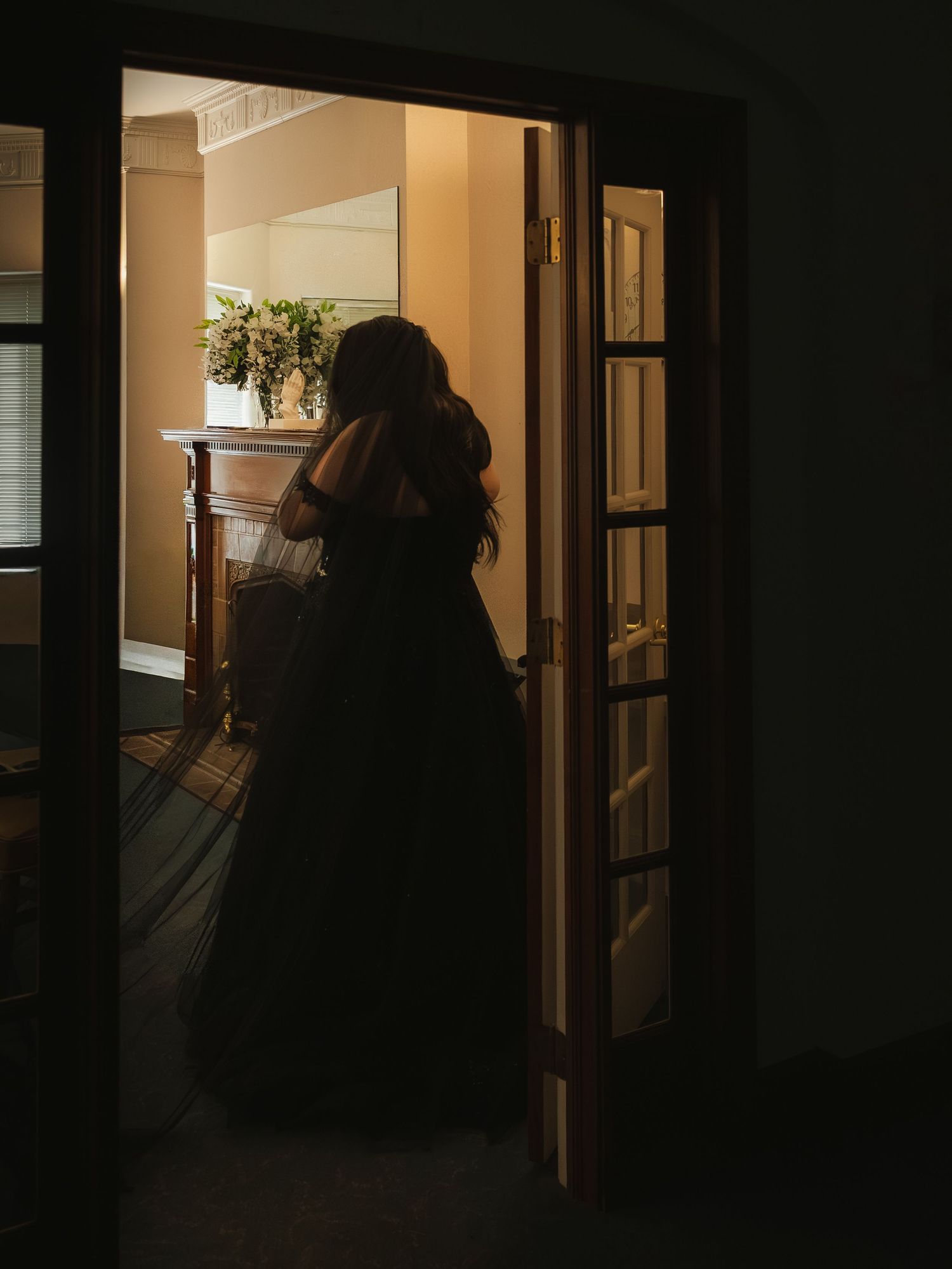 Bride in black wedding gown stands silhouetted in doorway of warmly lit bridal suite before ceremony.