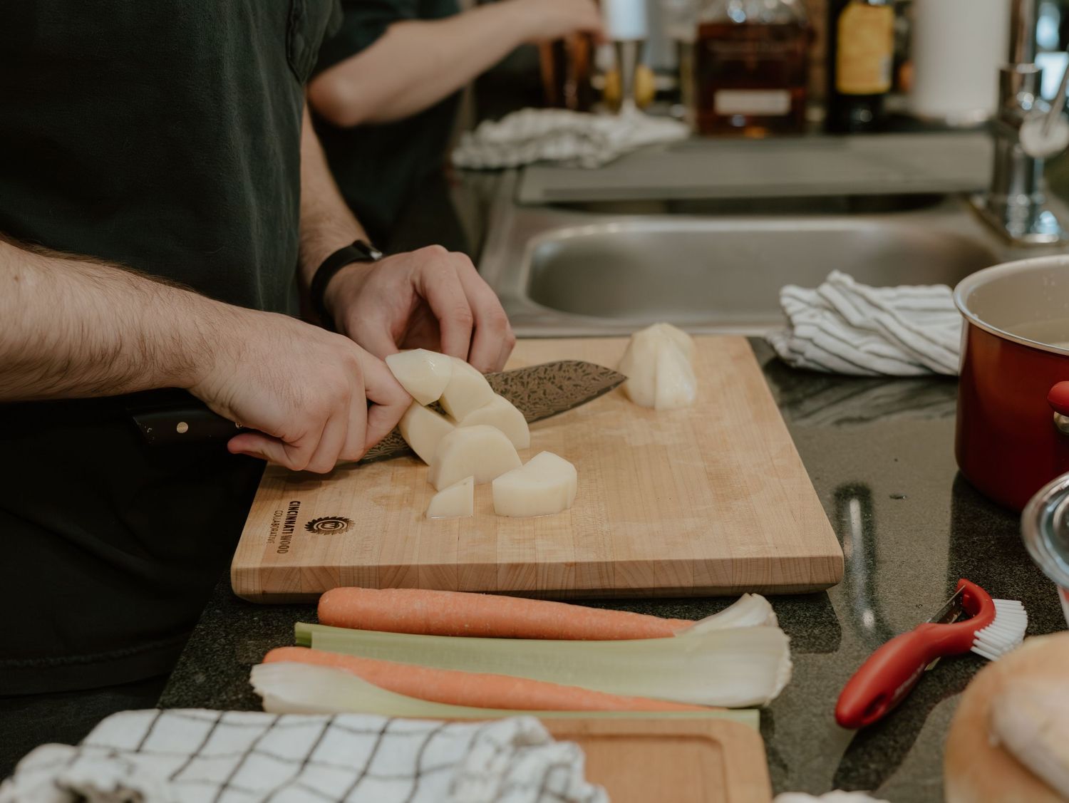 Someone chopping fresh ingredients on a wooden cutting board in a kitchen with carrots and kitchen tools nearby.