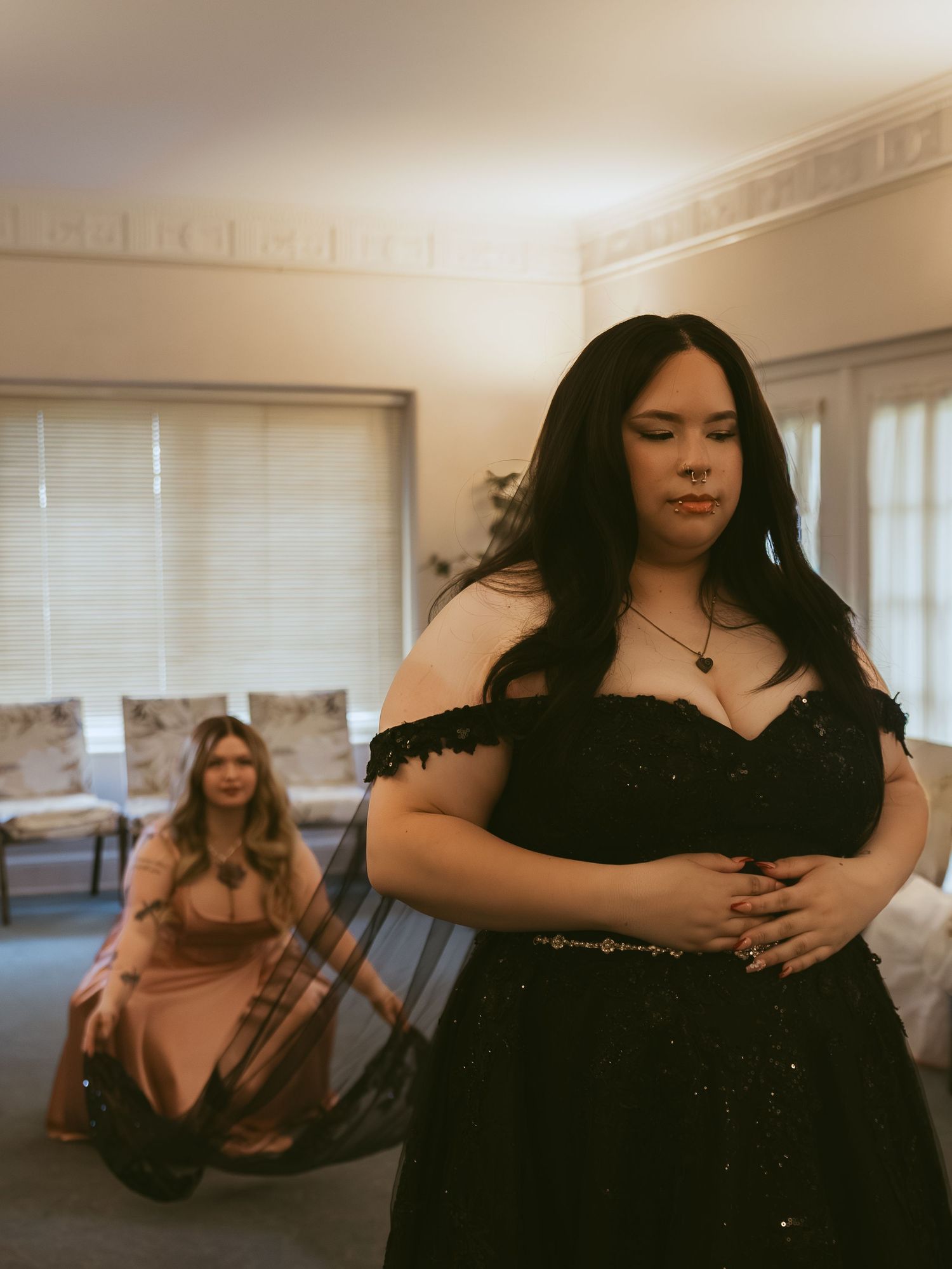 Two women in formal gowns, one in black off-shoulder dress foreground, one in pink seated behind in elegant room.