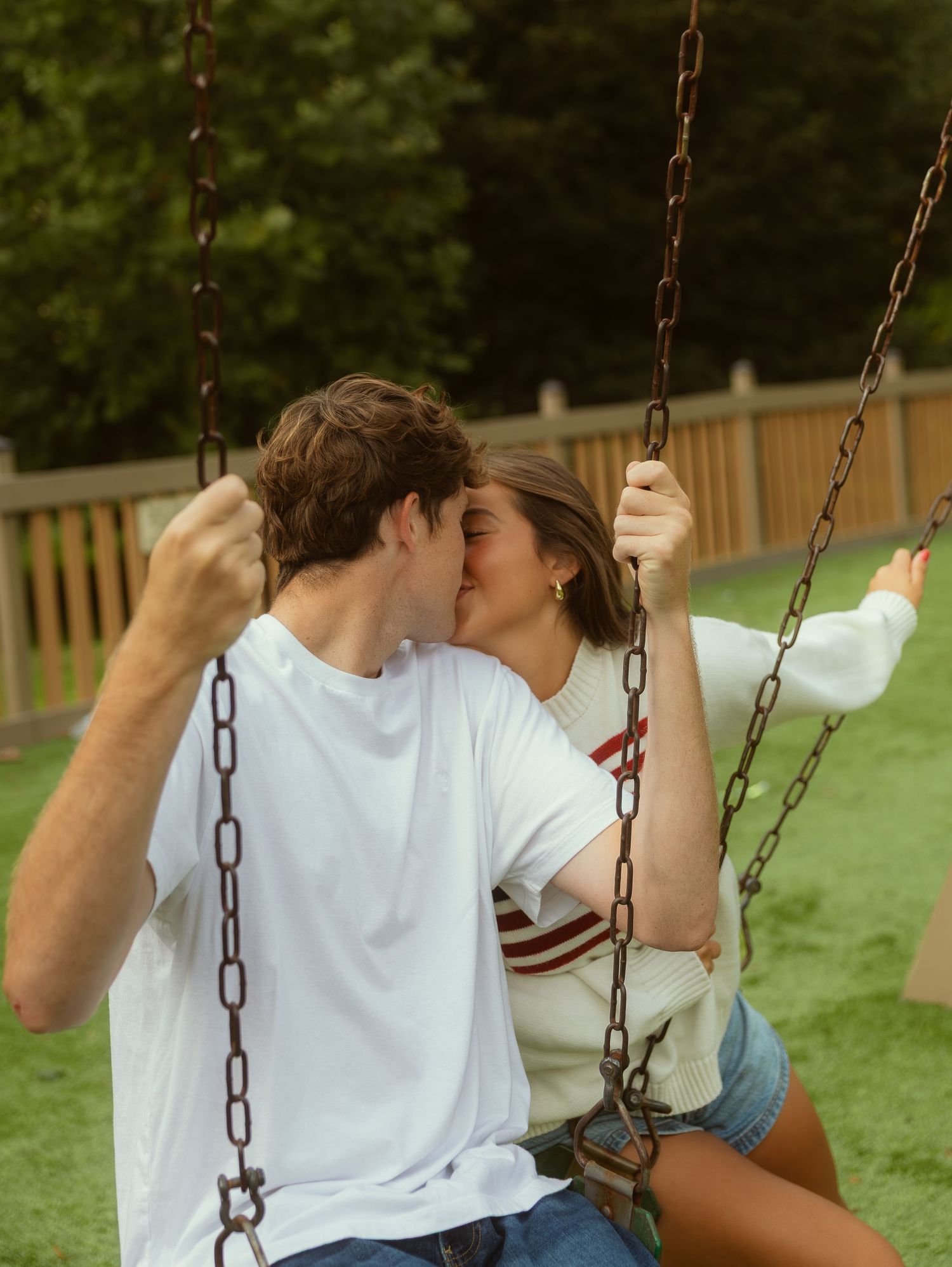 Young couple sharing a romantic kiss on a backyard swing set, surrounded by lush green trees and wooden fence.
