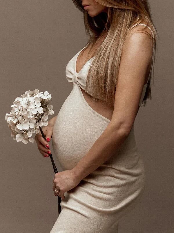 Pregnant woman in cream dress holds white hydrangea flowers against neutral backdrop in elegant maternity portrait.