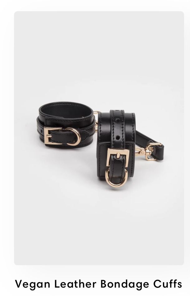 Black vegan leather bondage cuffs with gold-tone buckles and D-rings displayed on white background.