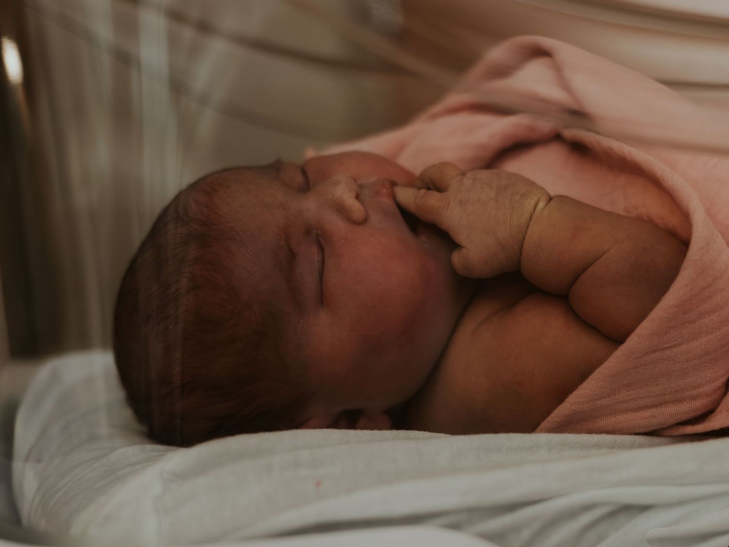 A peacefully sleeping newborn baby wrapped in a soft pink blanket rests on white bedding in warm lighting.