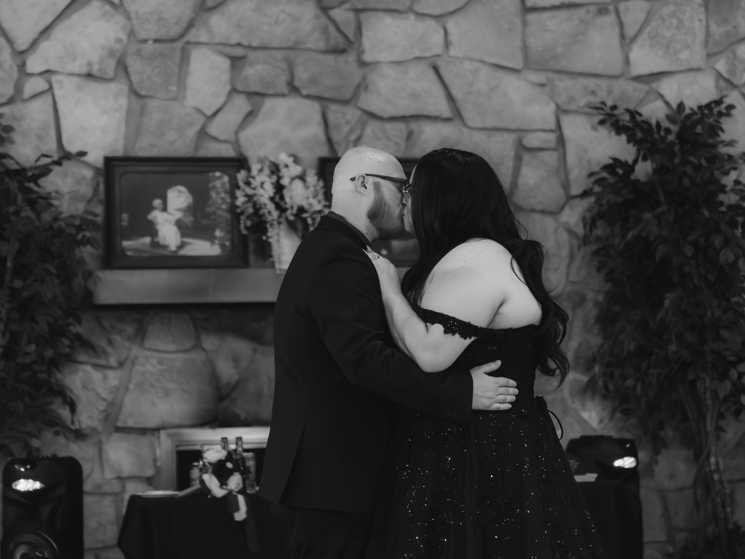Black and white photo of a couple sharing a kiss near a stone fireplace at a formal event.