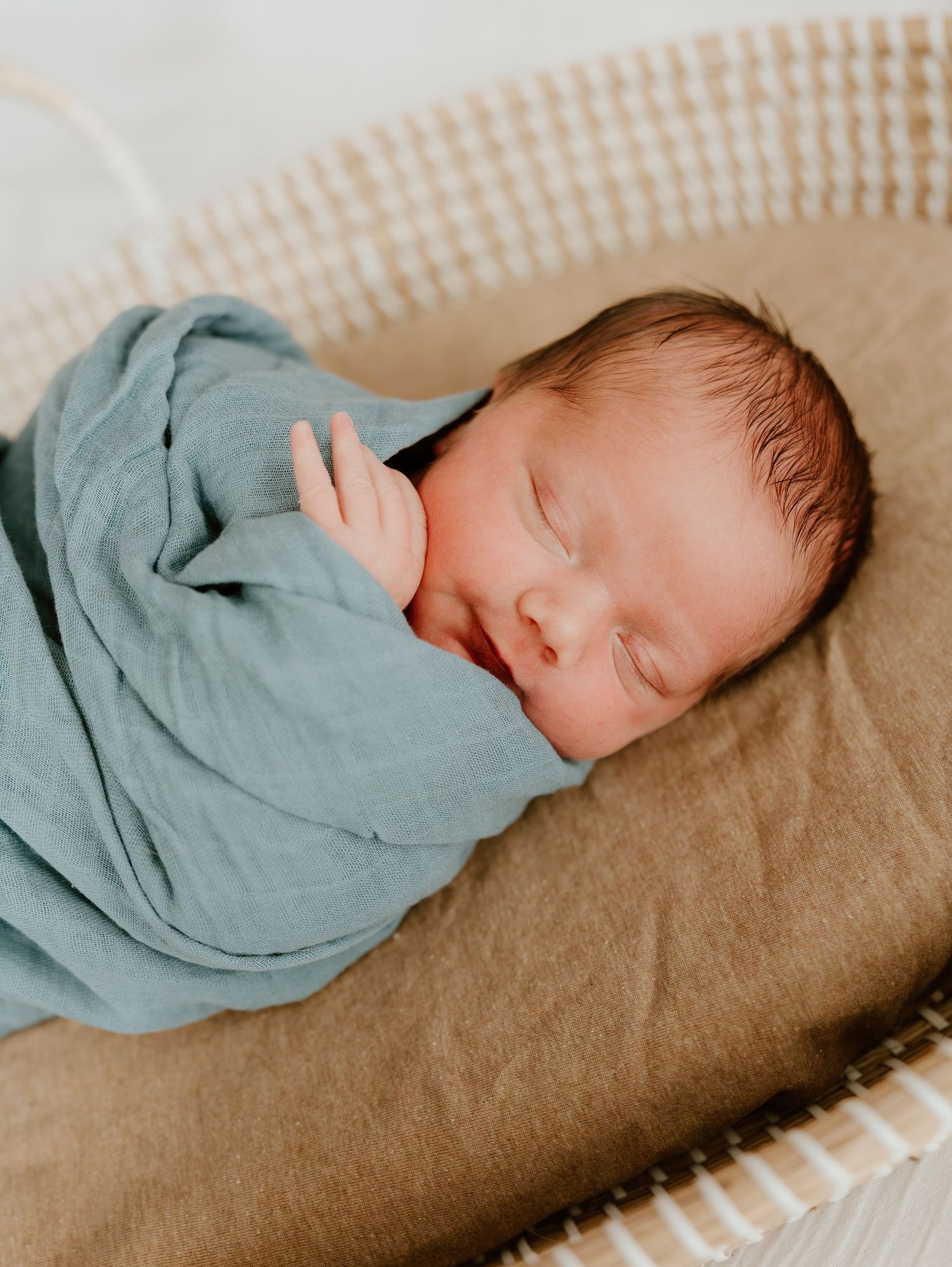 A sleeping newborn baby wrapped in a soft teal swaddle blanket lies peacefully on a beige textured surface.