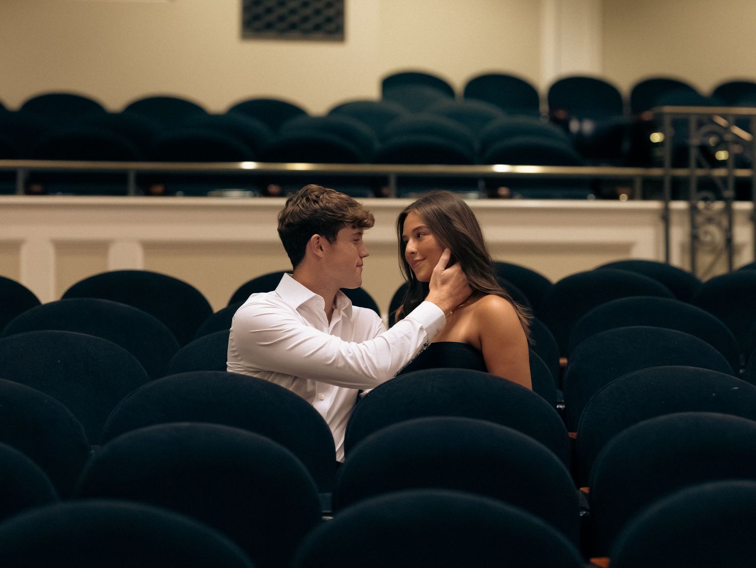 Two people share an intimate moment in an empty theater with rows of dark blue seats.