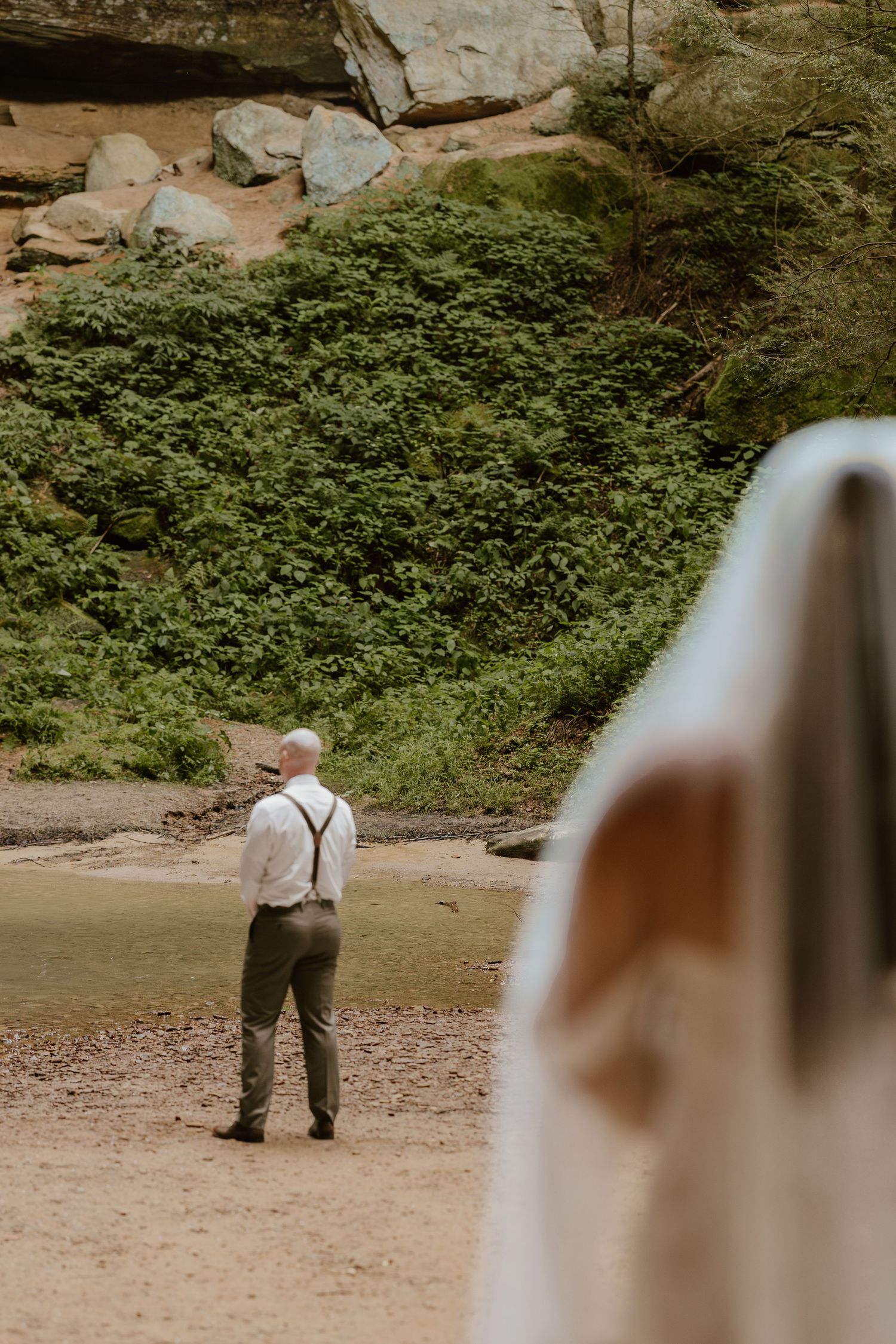 A person in light clothing walks away on a sandy path surrounded by lush greenery and rocky cliffs.