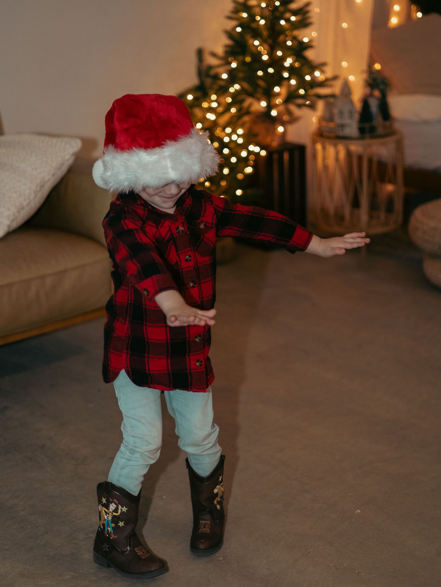 A small figure in a red plaid shirt and Santa hat dances in front of a lit Christmas tree in a cozy living room.