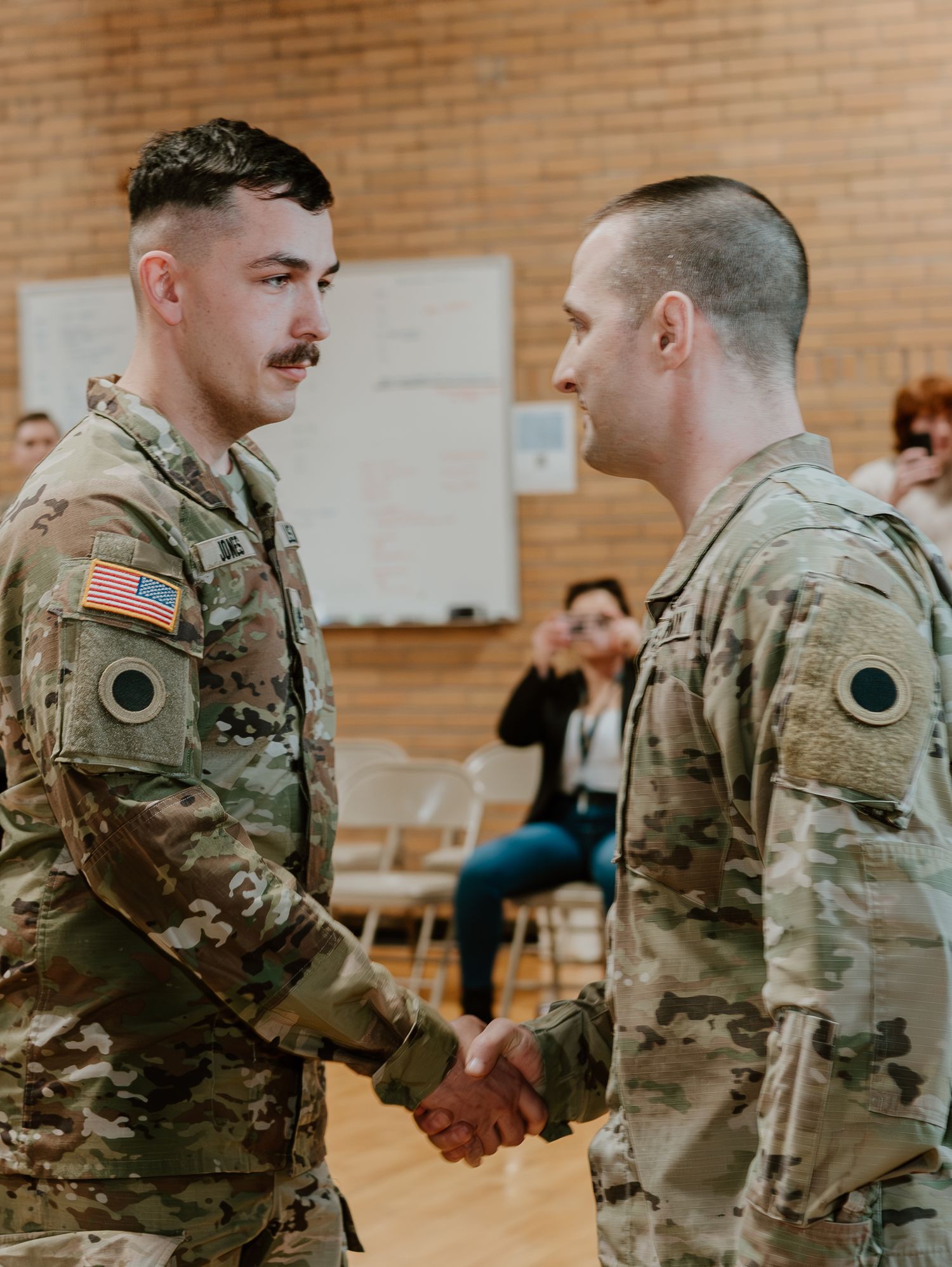 Two soldiers in camouflage uniforms shake hands in what appears to be a gymnasium with brick walls.