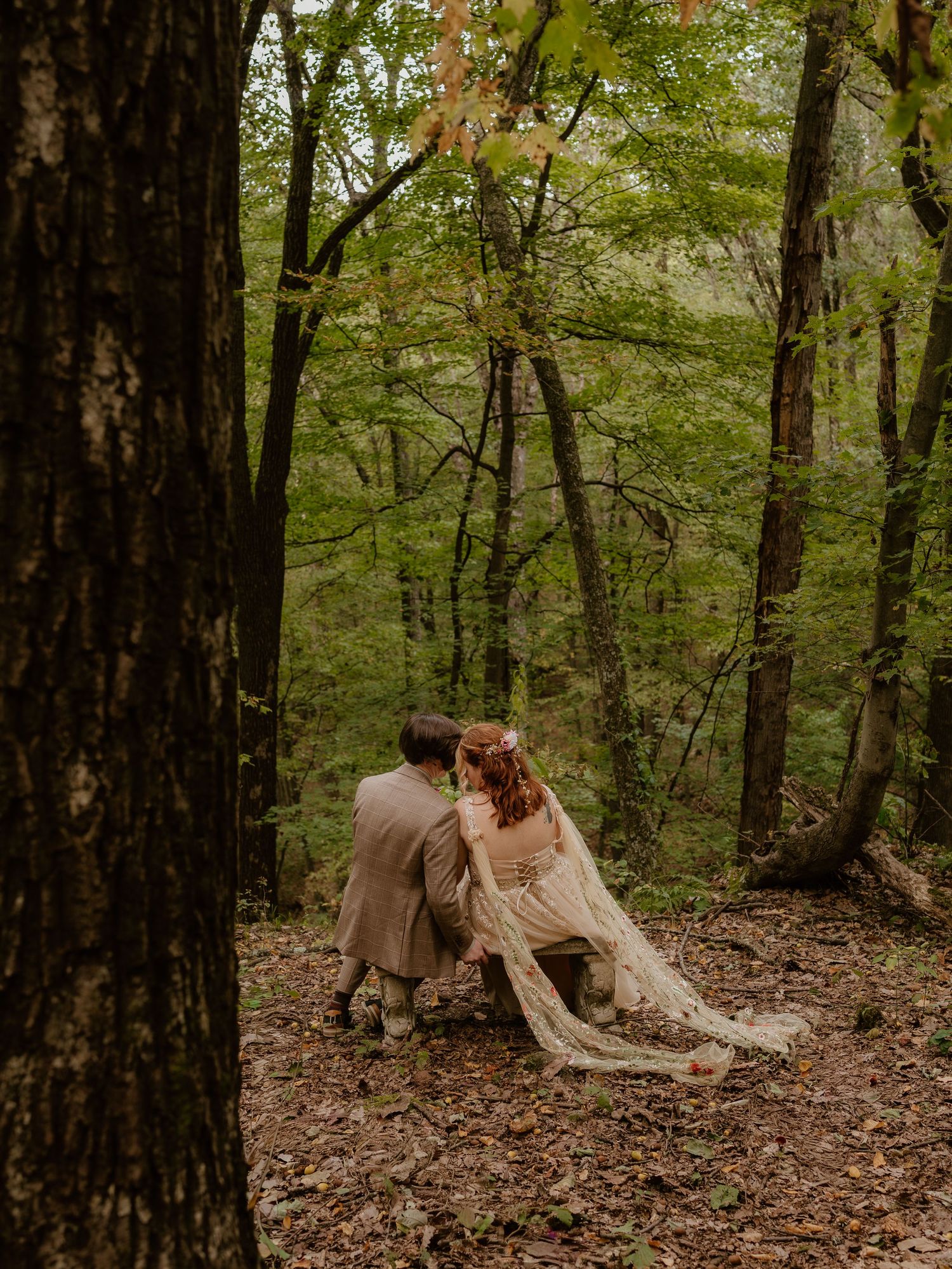 A couple in elegant attire lies together on autumn leaves in a serene forest setting during a romantic photoshoot.