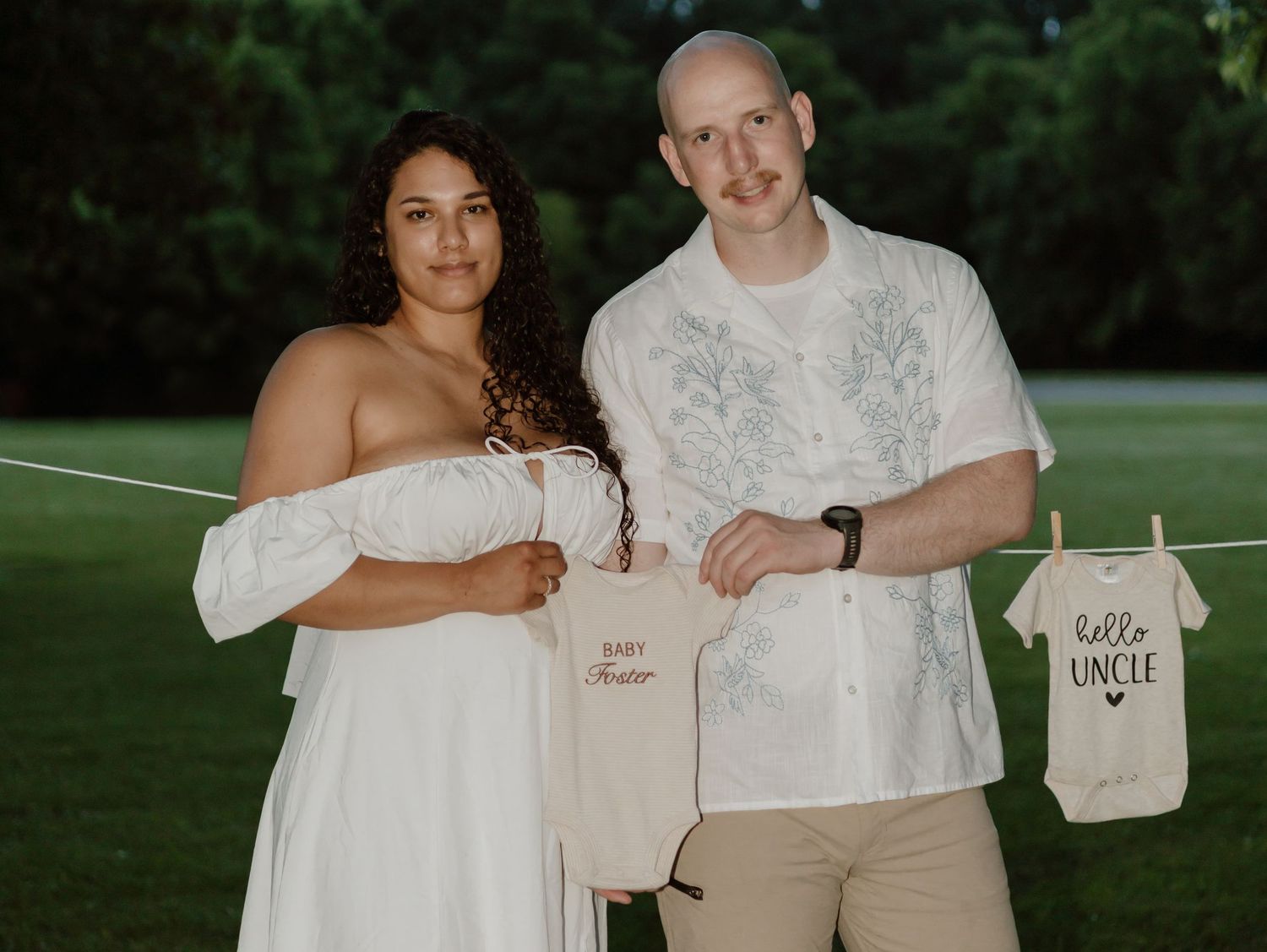 Couple holding baby onesies with text in an outdoor setting at sunset.